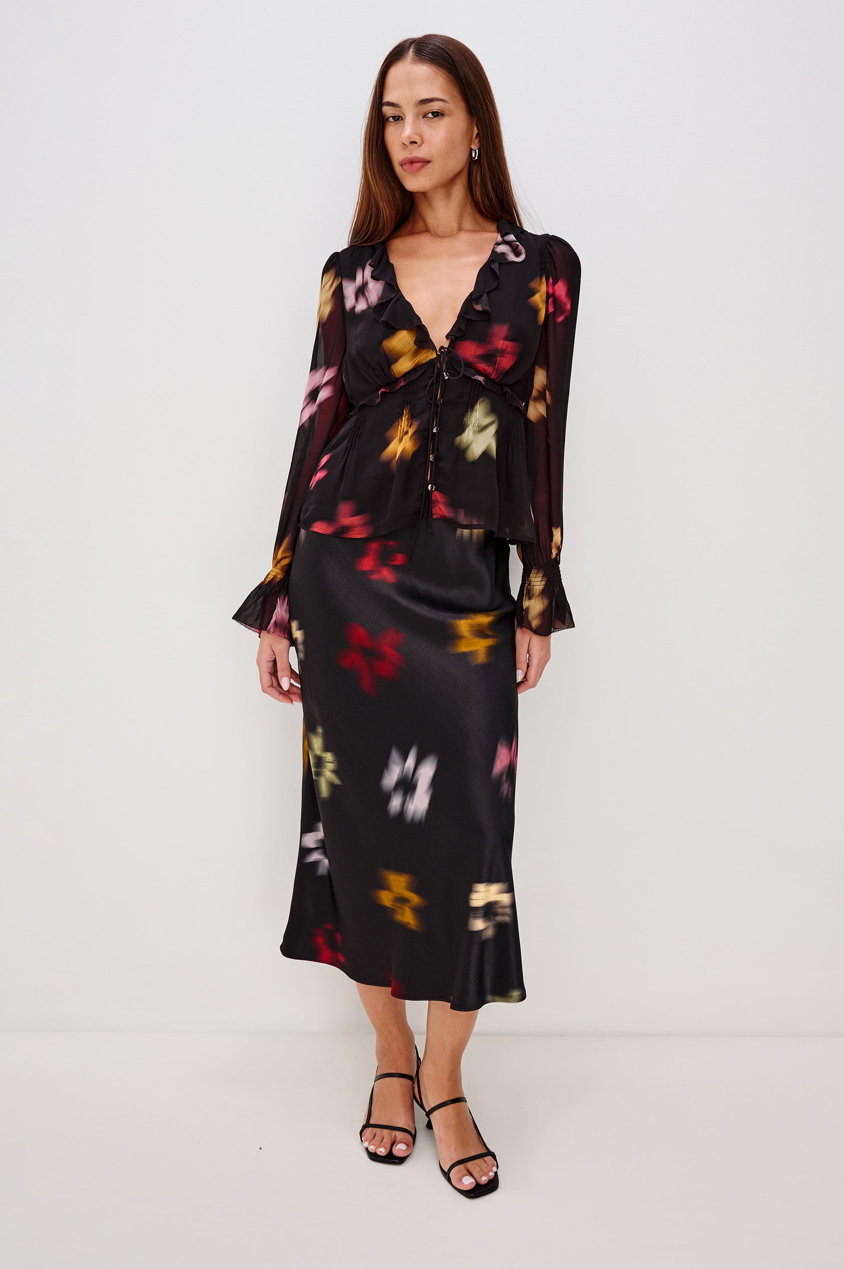 A lightweight chiffon blouse with a blurred floral print. The top features a deep V-neckline with cascading ruffles, button-front closures, and long sleeves finished with smocked bell cuffs. Full Body Front View Featured on Model.