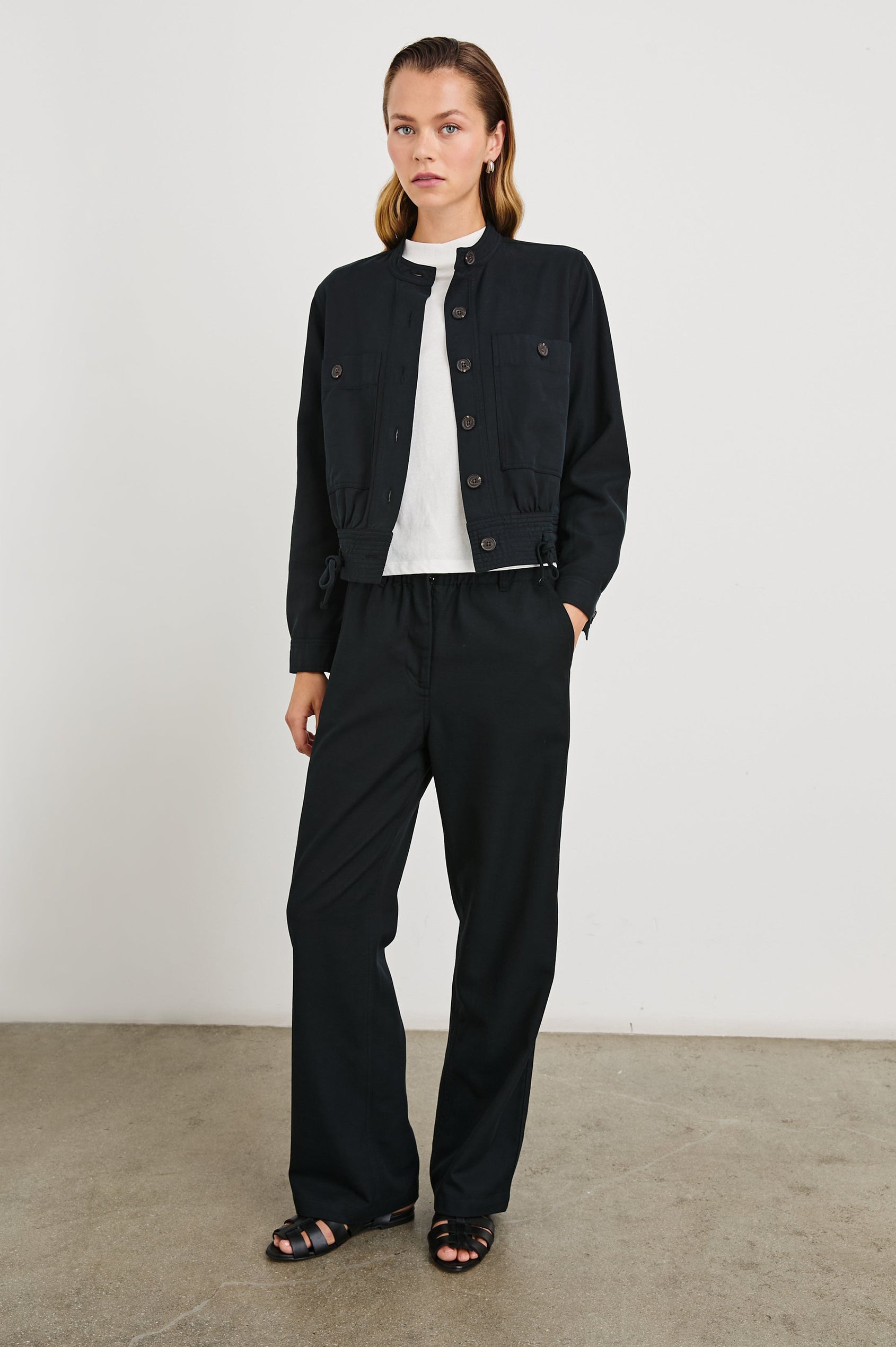 Alma Jacket with Button Closure, Mandarin Collar, Two Large Chest Button Patch Pockets and a Drawcord at the Hem in Black Colorway - Front Full Body View Featured on Model