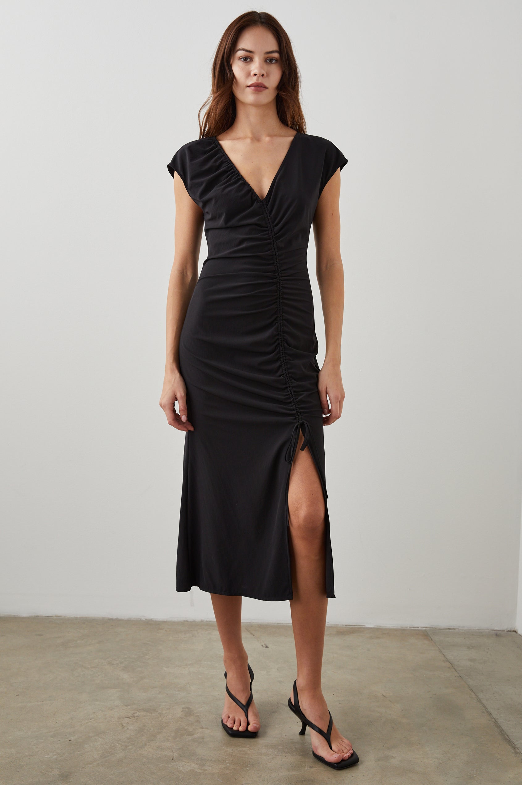 Auren Midi Dress with Cap Sleeves, a Deep V Neck, a Mid-Thigh Side Slit and Ruching from the V Neck to the Slit with a Drawcord at the Slit in Black Colorway - Front Full Body View Featured on Model