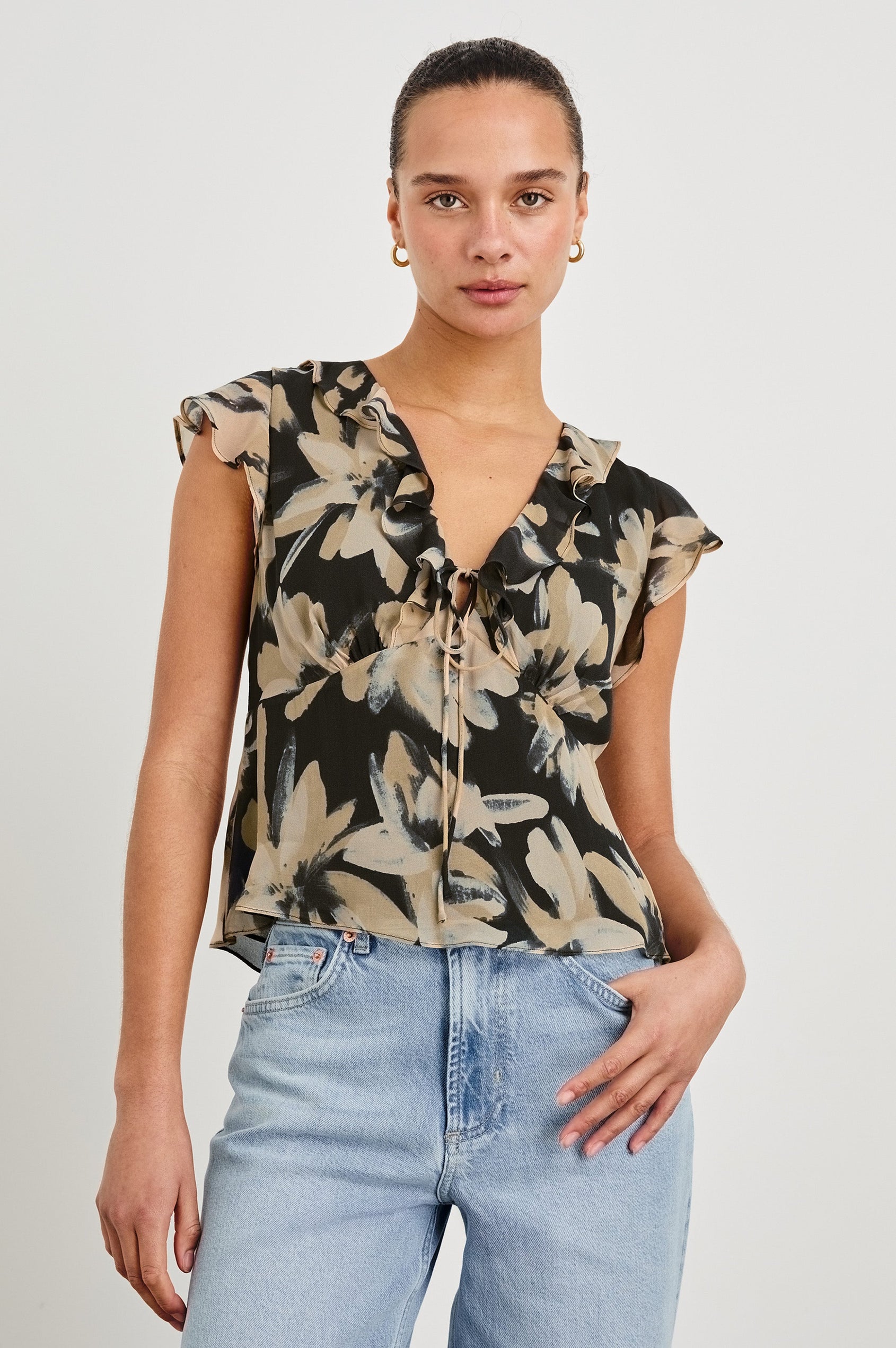 Model wearing our Carmine blouse, a chiffon top with flutter sleeves, ruffles at the v-neckline, a front tie, and empire waist silhouette. Styled with light washed denim pants.