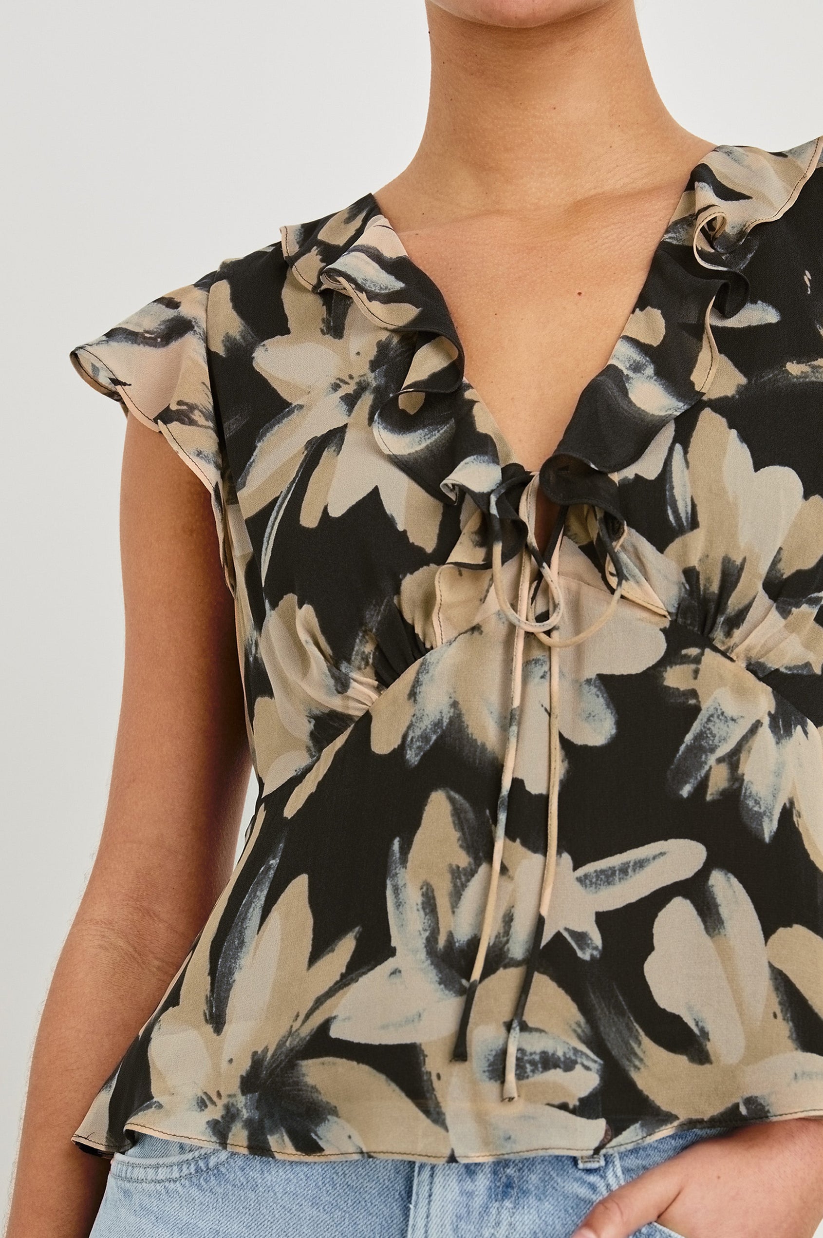 Detail view of model wearing our Carmine blouse, a chiffon top with flutter sleeves, ruffles at the v-neckline, a front tie, and empire waist silhouette. Styled with light washed denim pants.