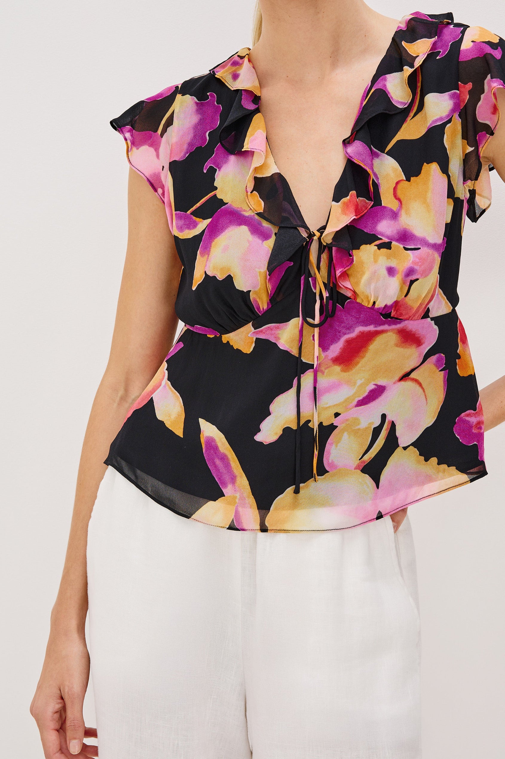 Carmine blouse, a chiffon top with ruffled cap sleeves, ruffles at the v-neckline, a front tie, and empire waist silhouette in Dusk Orchid Colorway. Front Detail View Shown on Model.