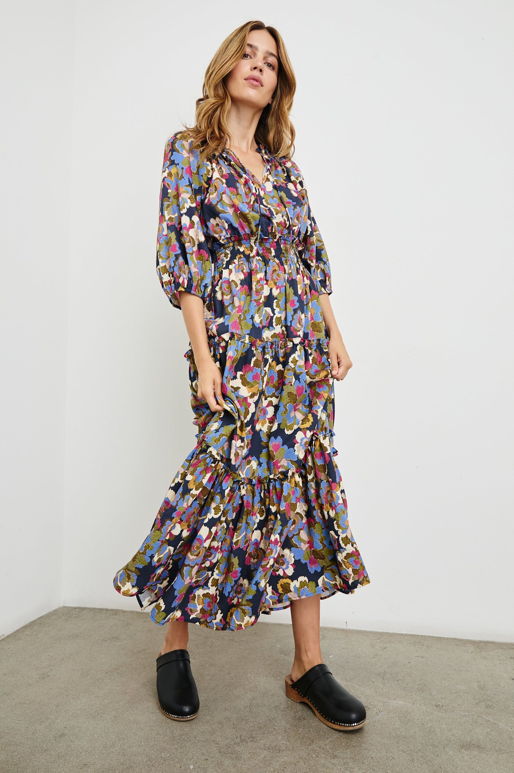 Caterine Maxi Dress with Mid-length Sleeves, a V neck, a Shirred Collar with a tie, a Smocked Waist, and a Tiered Skirt in Navy Wildflower Colorway, flowers in creams, pinks, greens and blues on Navy - Front Full Body View Featured on Model