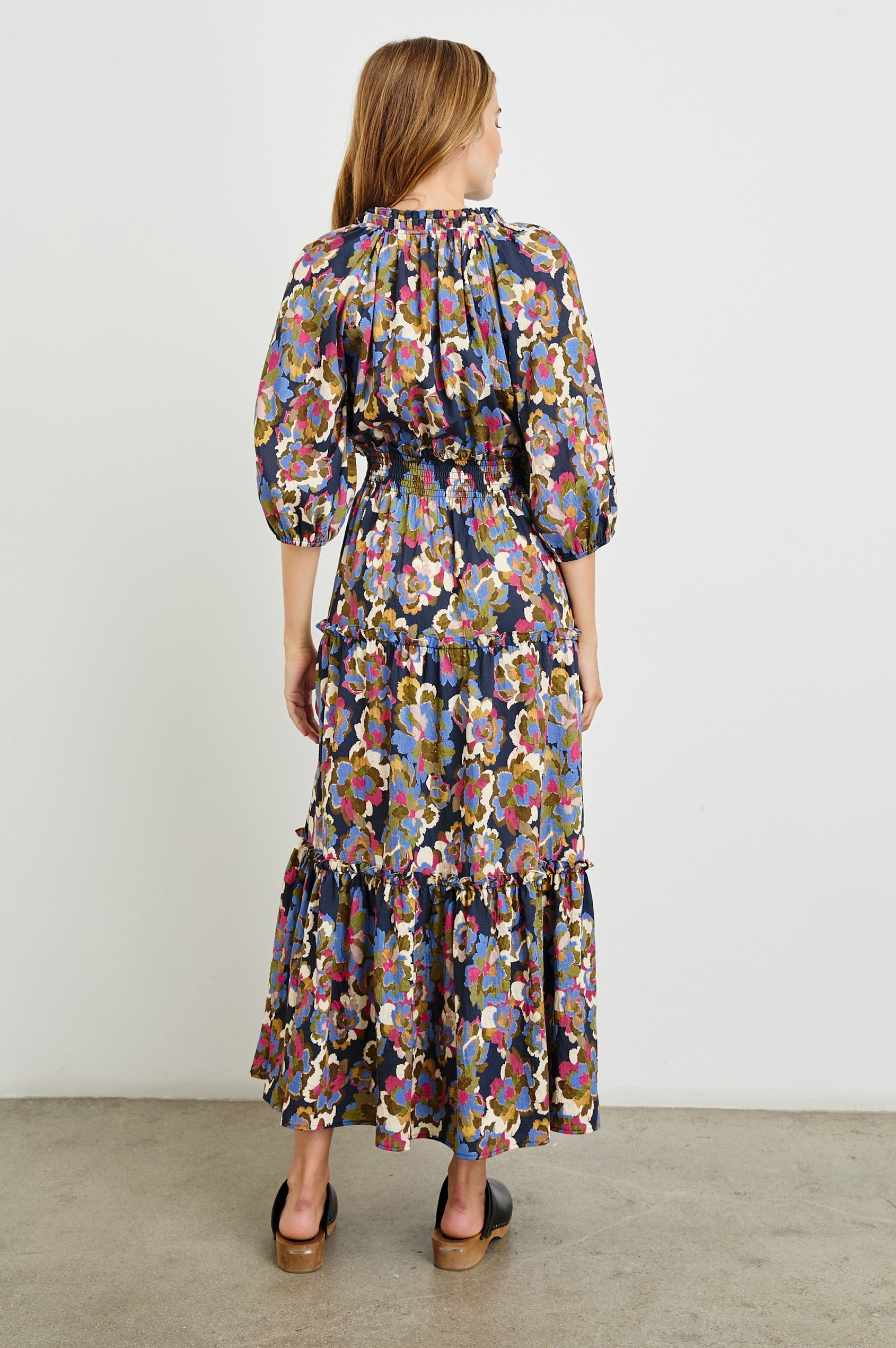 Caterine Maxi Dress with Mid-length Sleeves, a V neck, a Shirred Collar with a tie, a Smocked Waist, and a Tiered Skirt in Navy Wildflower Colorway, flowers in creams, pinks, greens and blues on Navy - Back Full Body View Featured on Model