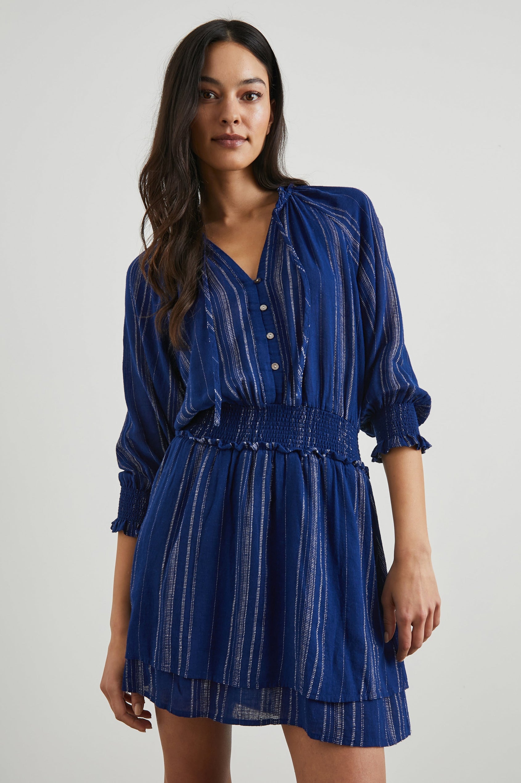 Crystal Mini Dress with Mid-Length Sleeves, Shirred Cuffs and Waist, a V Neck, Ties at the Collar, and Buttons down the Bodice in Noja Stripe Colorway, Thin White Vertical Stripes on Blue - Front View Featured on Model