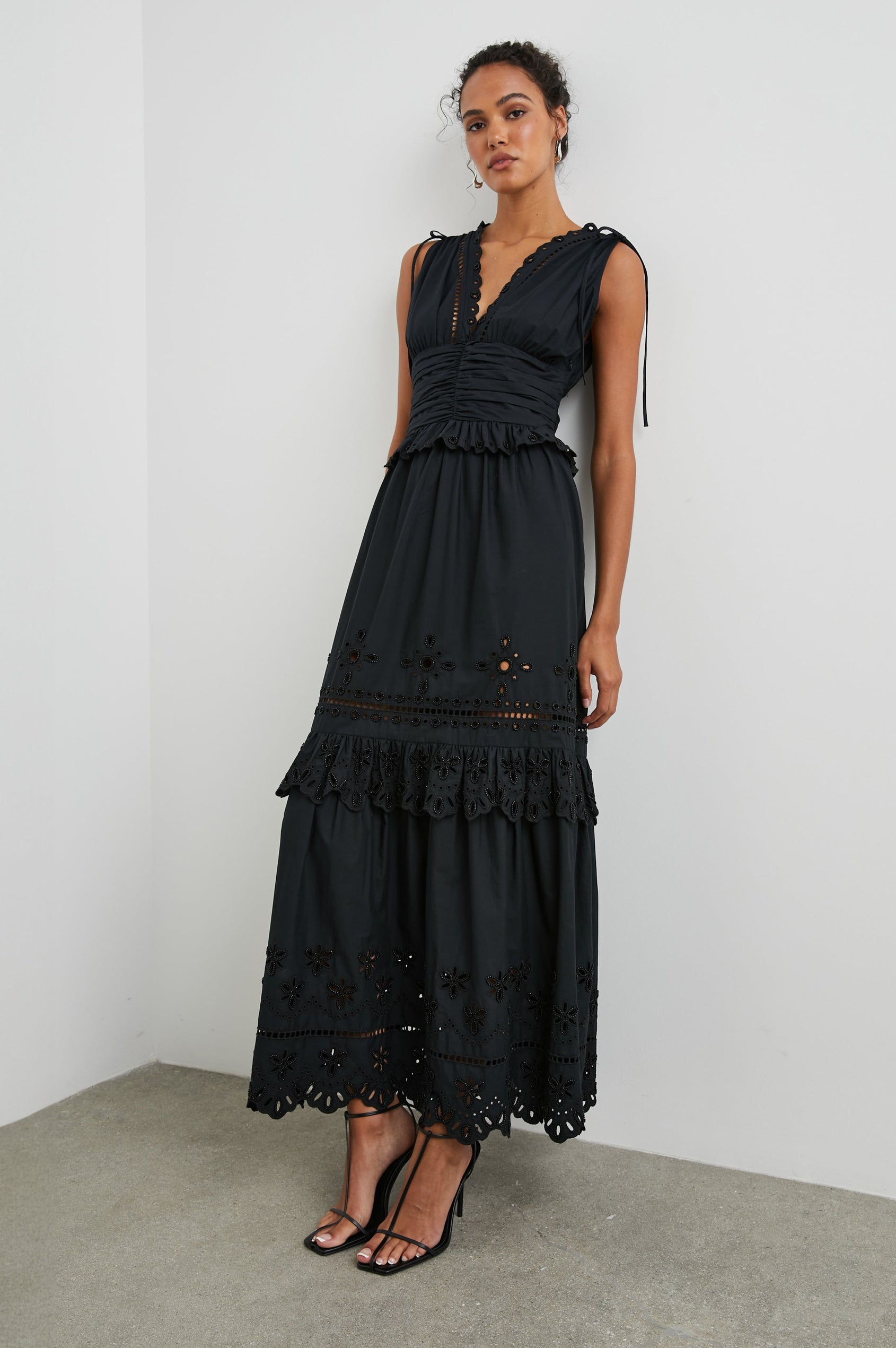 Esmeralda Sleeveless Maxi Dress with a Deep V Neck, a Ruched Waistband, Drawstrings Across the Shoulder Straps, Scalloped Trim with Cutouts on the Bodice, a Tiered Skirt, and an Eyelet Design along the Hem in Black Colorway - Front Full Body View Featured on Model