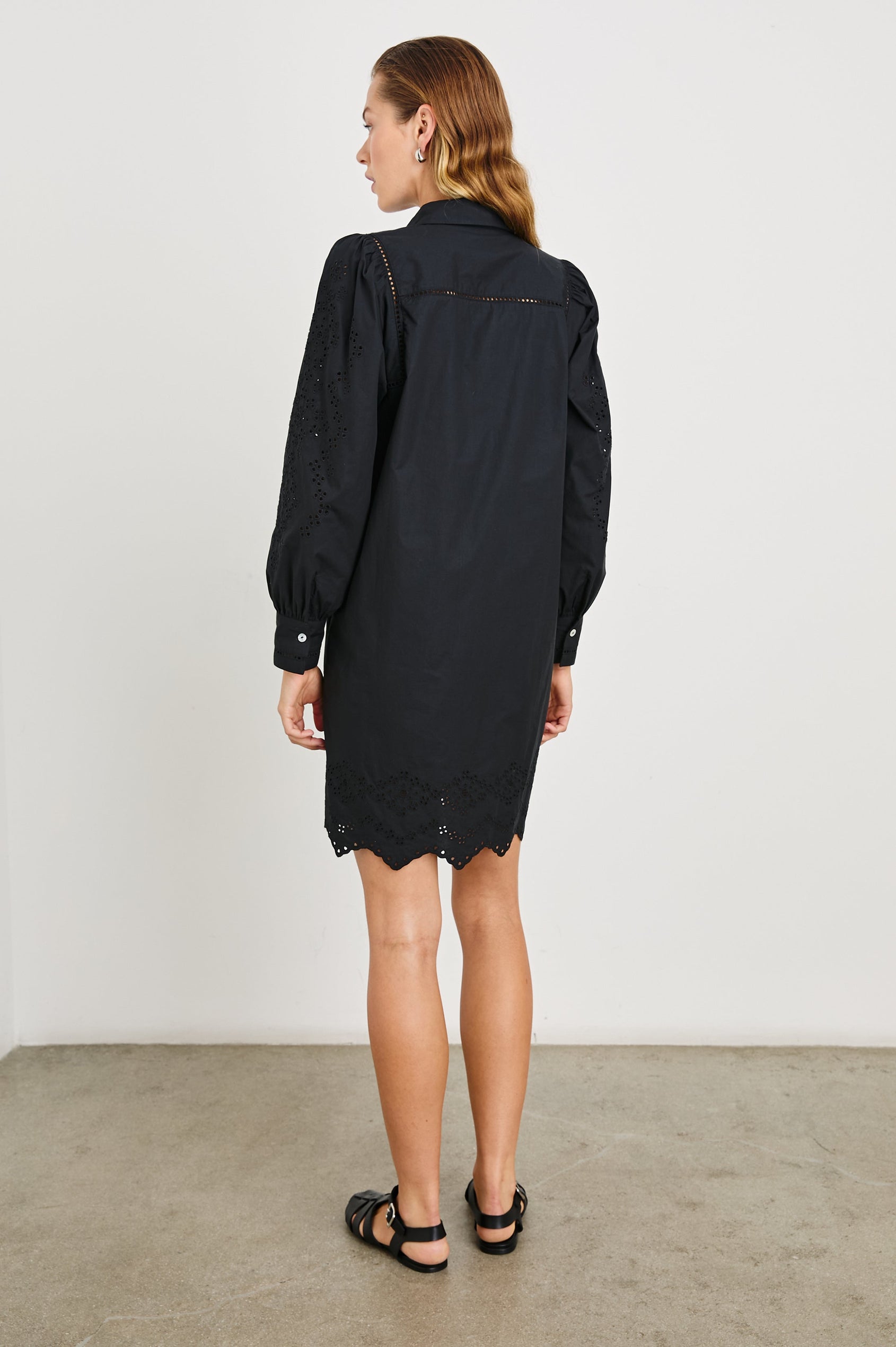 Fernanda Long-Sleeve Mini Dress with Button Cuffs, a Collar, a Placket with Buttons, a Scalloped Hem, a Floral Eyelet Design on the Sleeves and a Floral and Geometric Eyelet Design on the Hem in Black Eyelet Colorway - Back Full Body View Featured on Model