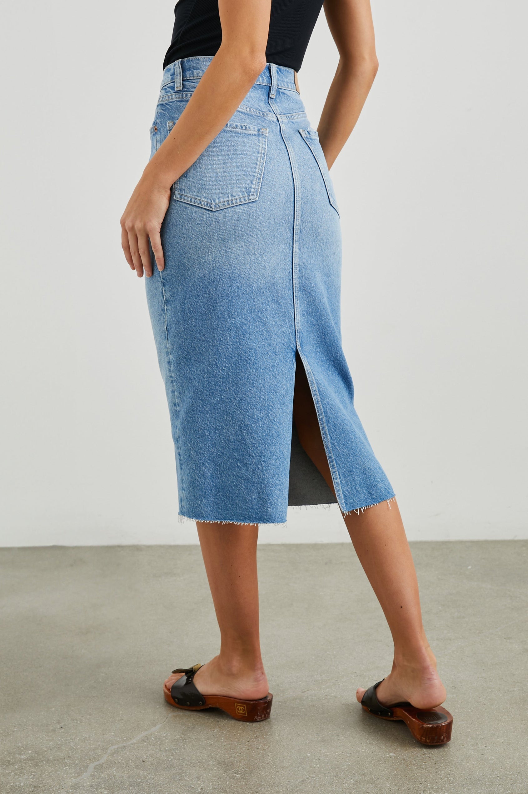 Highland Denim Midi Skirt with a Raw Hem, a Back Vent, Additional Front Seams, Belt Loops, Two Front Pockets plus a Coin Pocket and Two Back Patch Pockets in Baja Blue Colorway - Back View Featured on Model