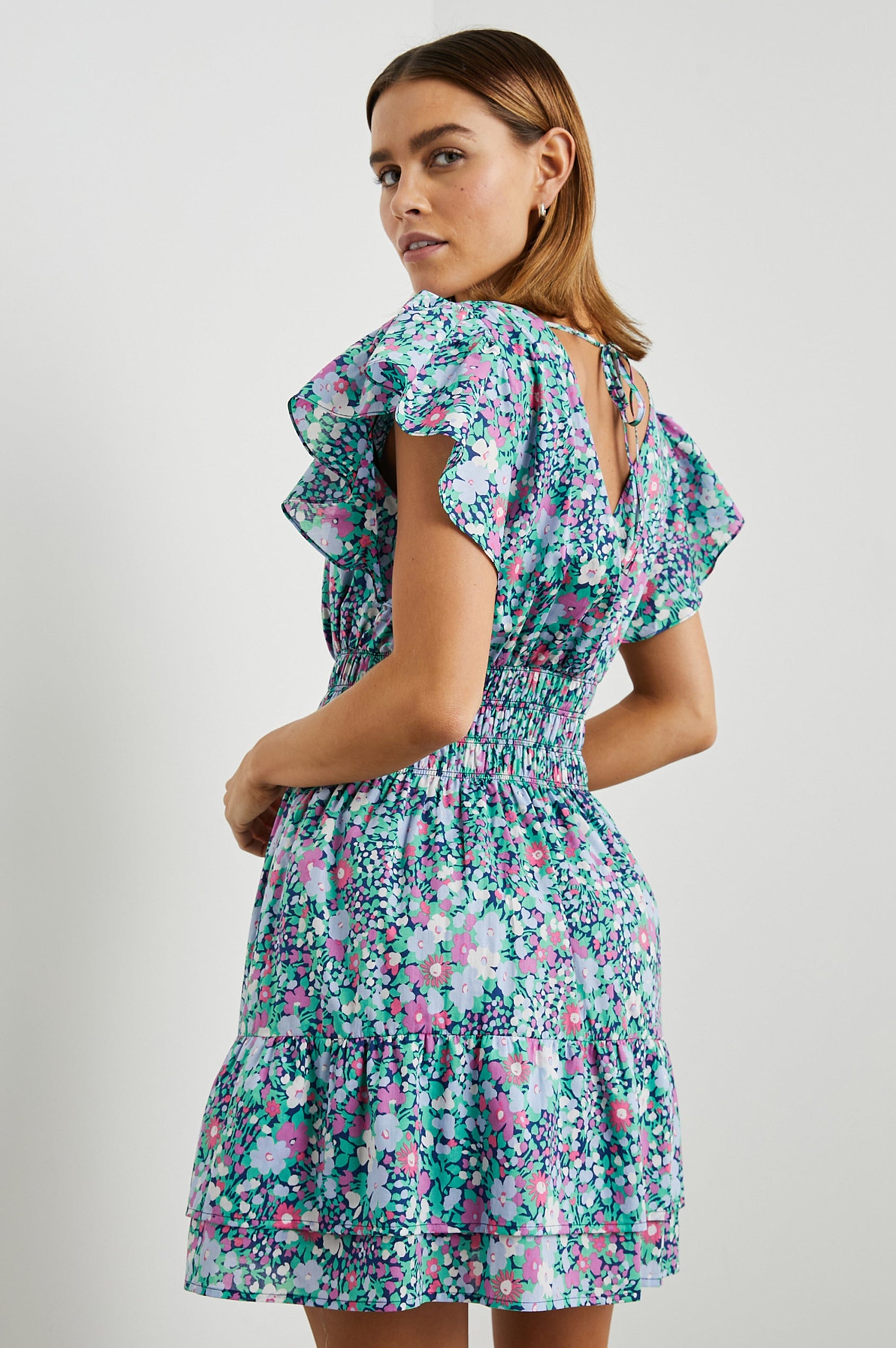 Kimora Above-Knee Dress with Flutter Sleeves, a V Neckline, a Cinched Waist and a Tiered Skirt in Navy Leilani Floral Colorway with flowers in pinks, purples and greens - Side View Featured on Model