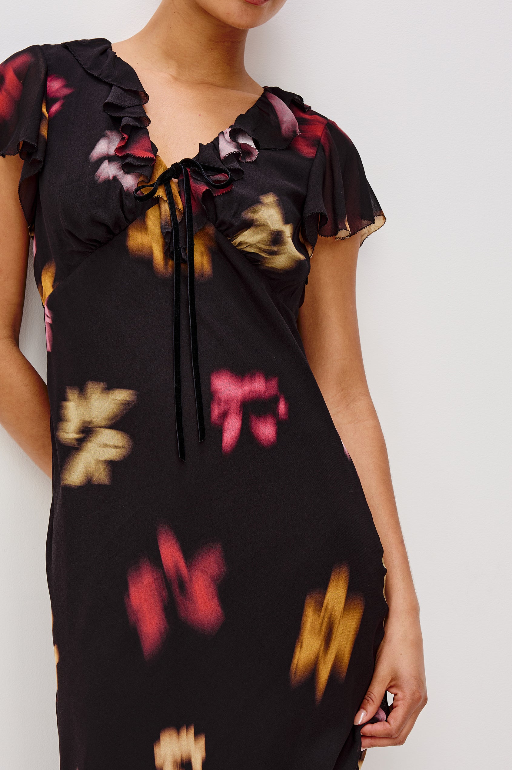 A chiffon midi dress with a blurred floral print. The dress has a V-neckline framed by cascading ruffles and a velvet bow, ruffle cap sleeves, and an empire waist that creates a graceful, elongated silhouette. Close Up View featured on Model.