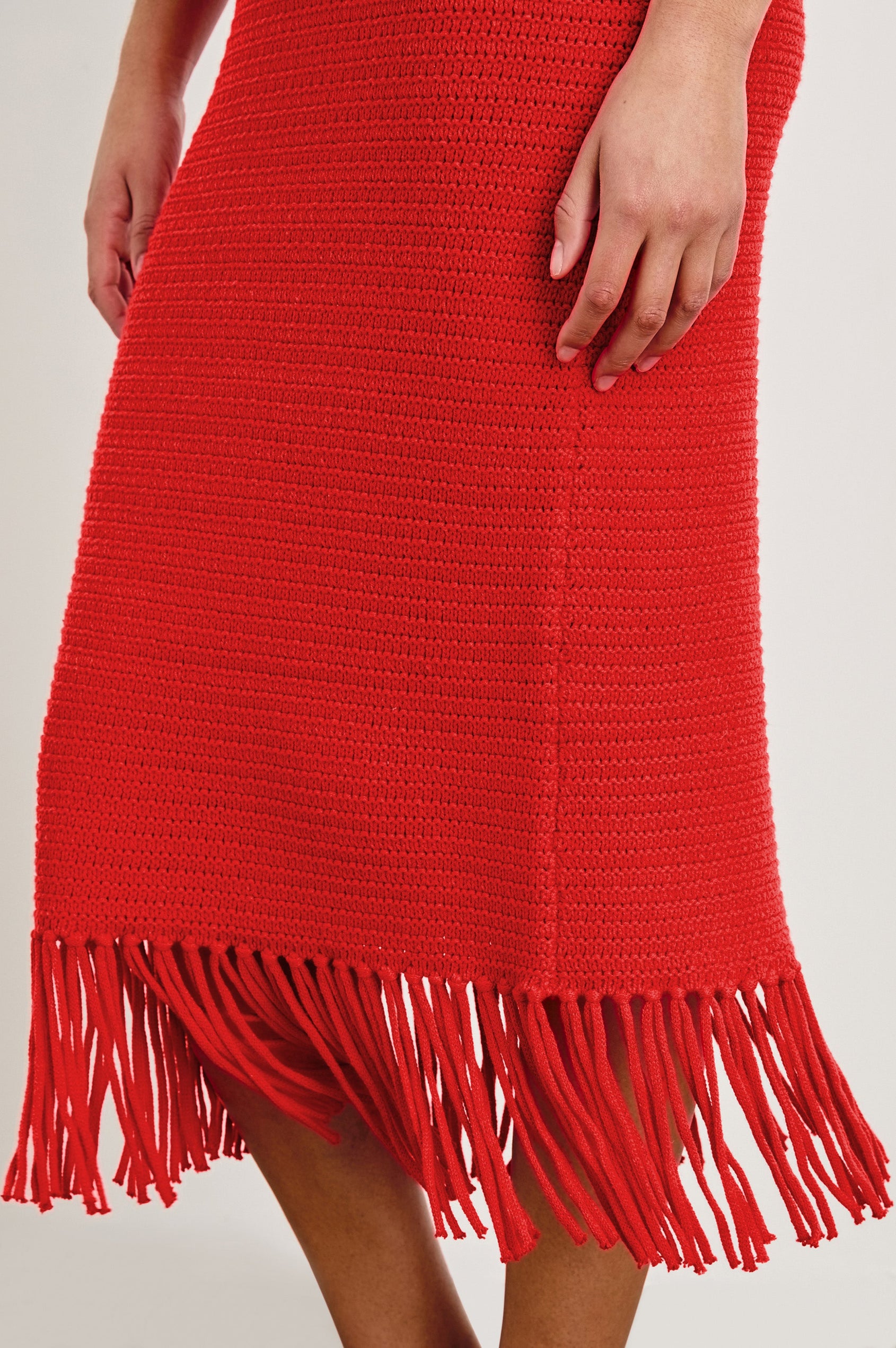 Detail view of female model wearing our sleeveless, Nia Midi Dress in color Cherry, featuring crochet structure and fringe details at the hem.