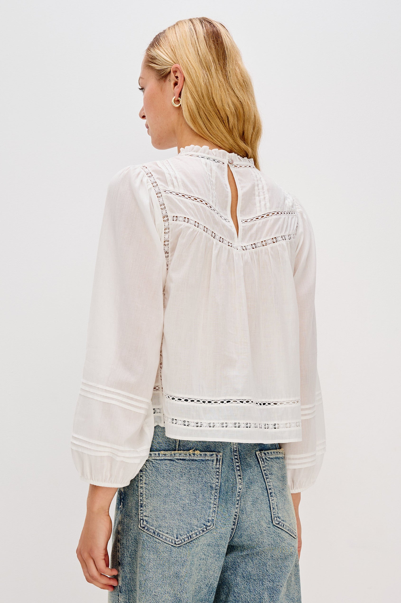Back view of female model wearing our Reyna Top in white. This is a long sleeve crewneck baby doll blouse, with ruffles at the collar.