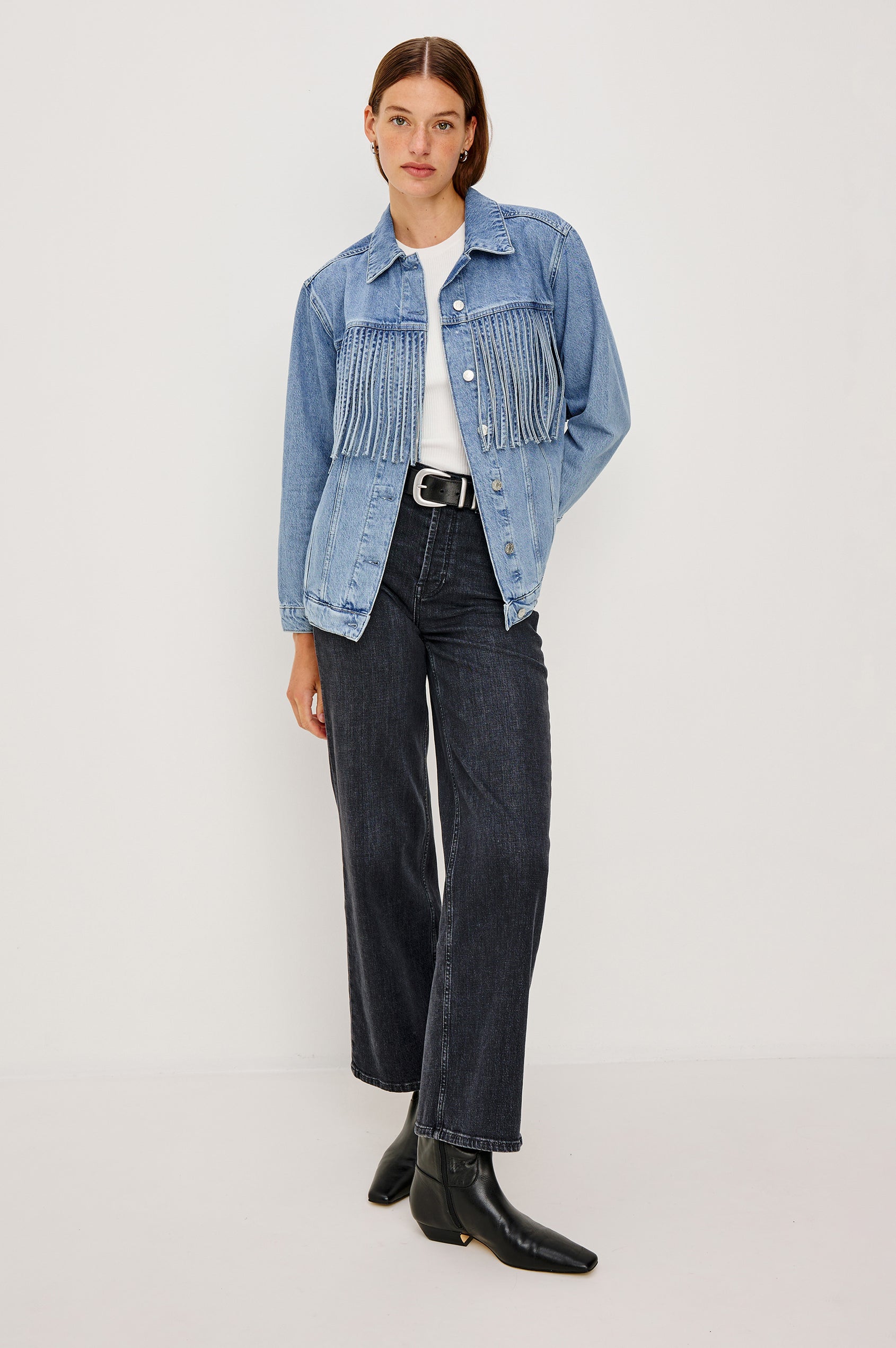 The Rodeo is button front collared denim jacket with side pockets and fringe along front yoke in Indigo Cowboy colorway, a medium blue wash. Front full body view featured on model.