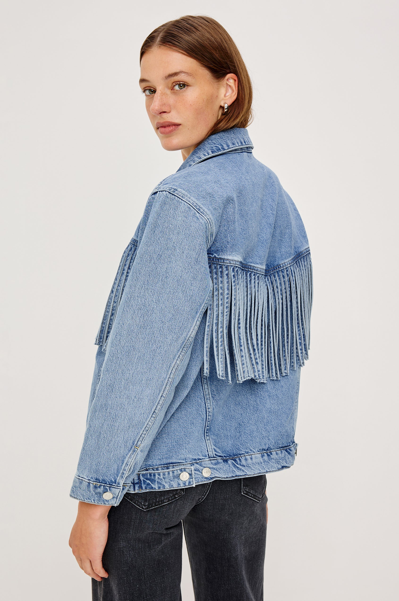 The Rodeo is button front collared denim jacket with side pockets and fringe along front yoke in Indigo Cowboy colorway, a medium blue wash. Back view featured on model.