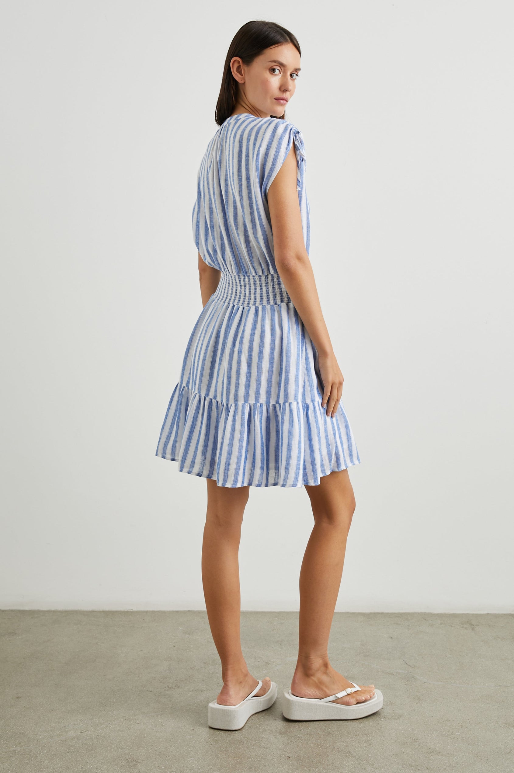 Samina Above-Knee Sleeveless Dress with drawstrings at the Sleeve Openings, Buttons along the Bodice, A Cinched Waist and a Tiered Skirt in Casablanca Stripe Colorway, with blue vertical stripes on white - Back Full Body View Featured on Model