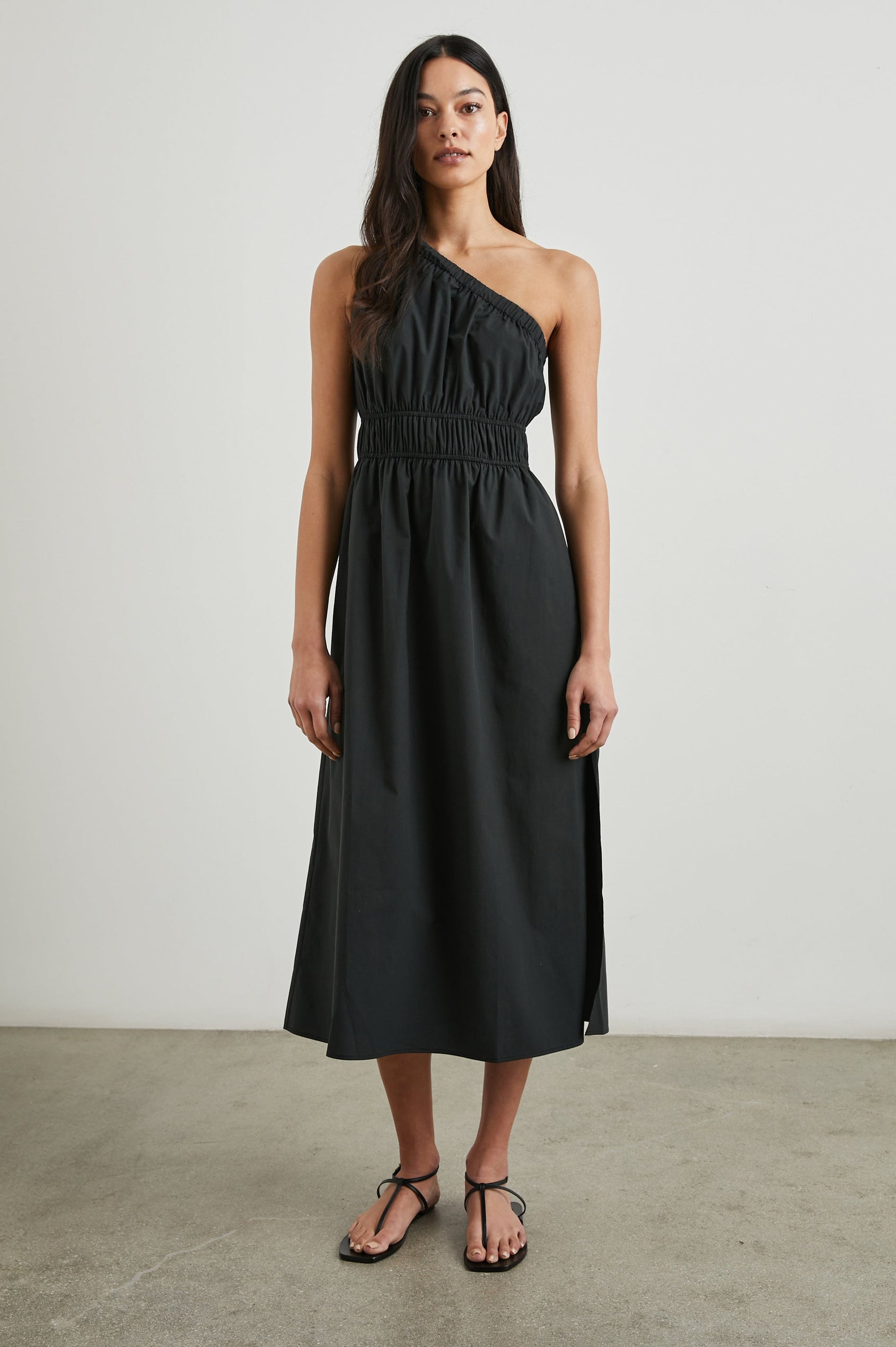 Selani Midi Dress With an Asymmetrical Neckline, Mid-Thigh Slide Slit and Cinched Waistband in Black Colorway - Front Full Body View Featured on Model