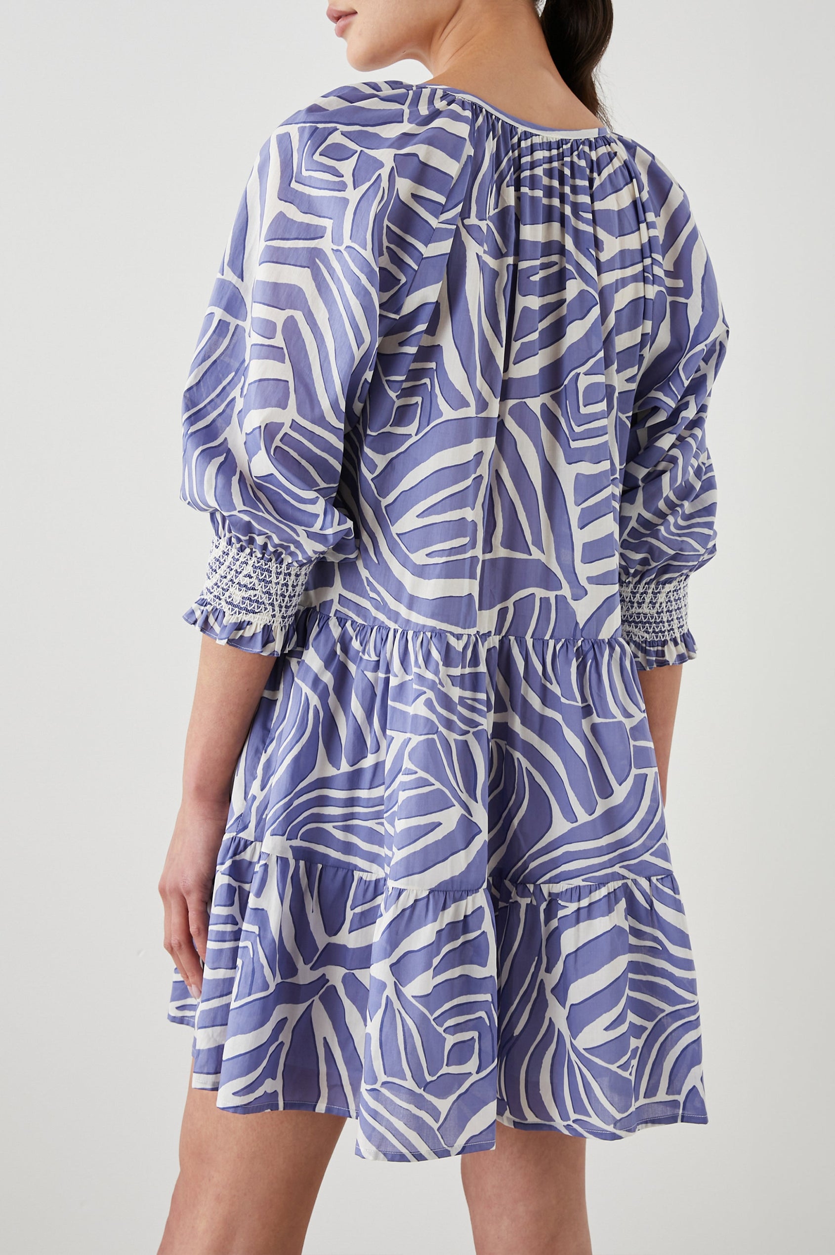 Sia Mini Dress with Three-Quarter Sleeves, Shirred Cuffs, a Tiered Skirt, and a V Neck with a Tie and Light Ruching in Island Waves Colorway, a Purple and White Print - Back View Featured on Model