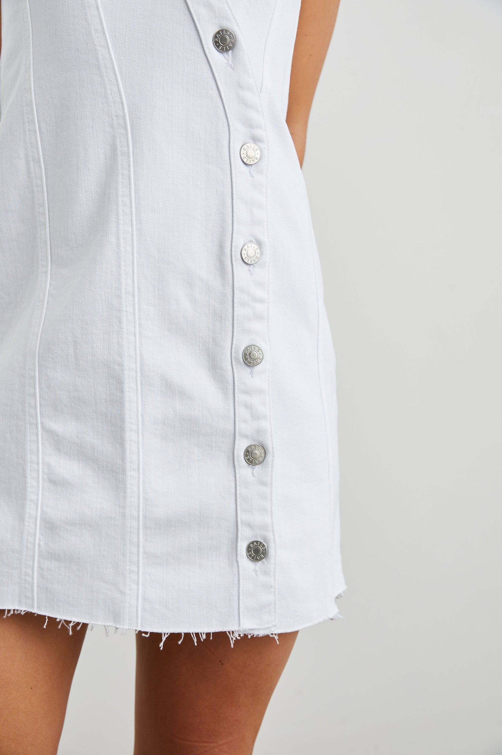 Strand Sleeveless Denim Mini Dress with a V Neck, a Raw Hem, Wide Shoulder Straps, a Row of Buttons that Extend Diagonally Across the Bodice and then Vertically Down the Skirt, Vertical Seams in Salt White Colorway - Front View Featured on Model