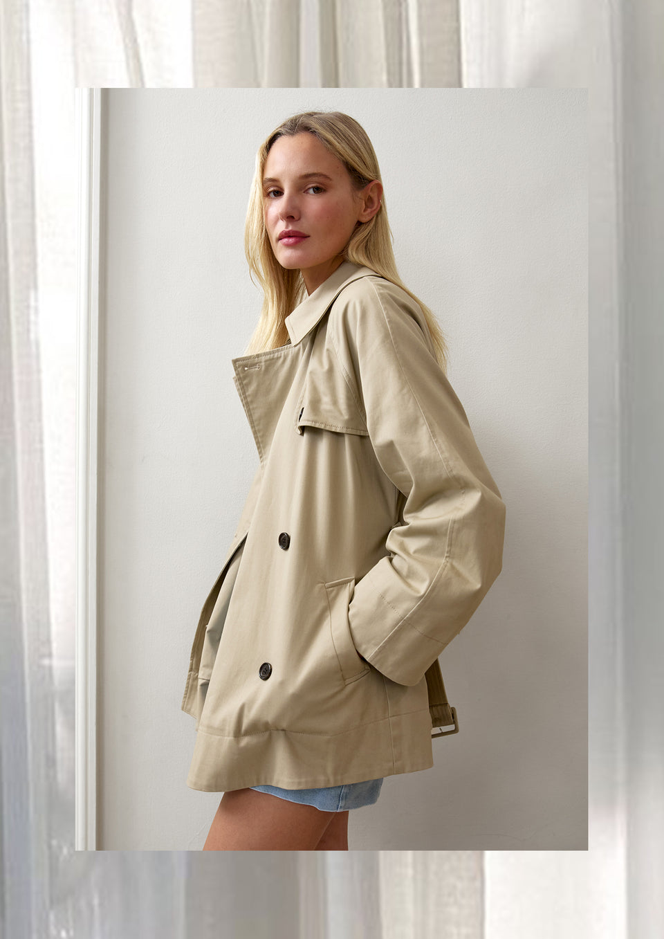 FEMALE MODEL STANDING TO THE SIDE WEARING LUCIEN JACKET IN COLOR TAUPE (SHOPT TRENCH JACKET IN LIGHT TAN COLOR)