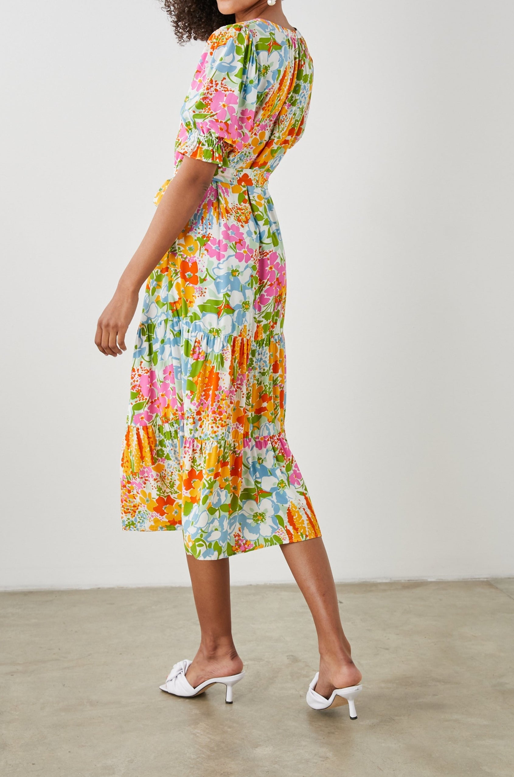 Eliana Midi Dress with Elbow-Length Sleeves with Shirred Cuffs, a V Neck, Buttons Down the Bodice, a Fabric Tie at the Waist and a Tiered Skirt in Day Garden Colorway, a Floral Pattern in Blue, Pink and Oranges on White - Back Full Body View Featured on Model