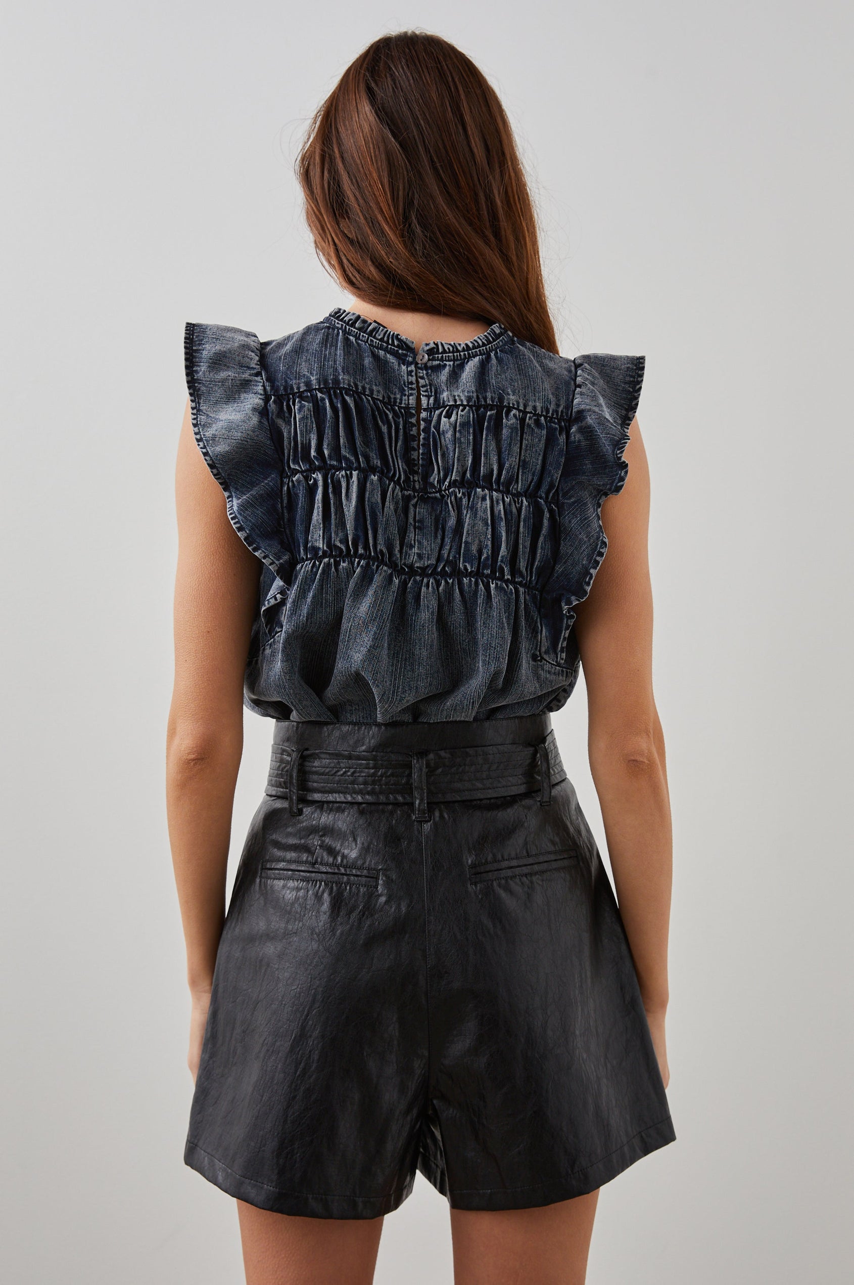 Filomena Short Sleeve Denim Top with Flutter Sleeves, a Ruffled Neck, a Shirred Center Bodice, Lace Inserts and a Buttoned Keyhole at the Back in Ink Wash Colorway - Back Tucked View Featured on Model