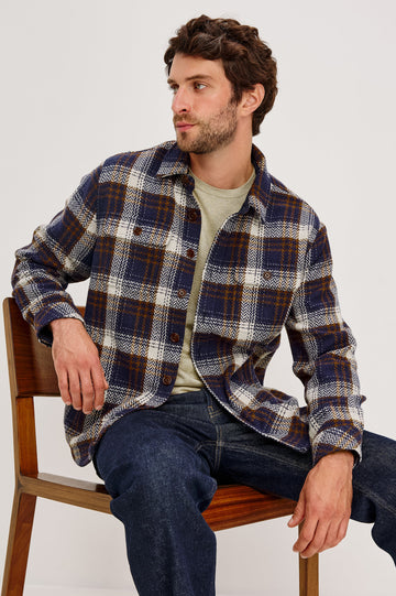 The Berkshire is a plaid shirt jacket with two chest pockets button front and collar in Navy Umber Cream colorway. Front seated view featured on model.