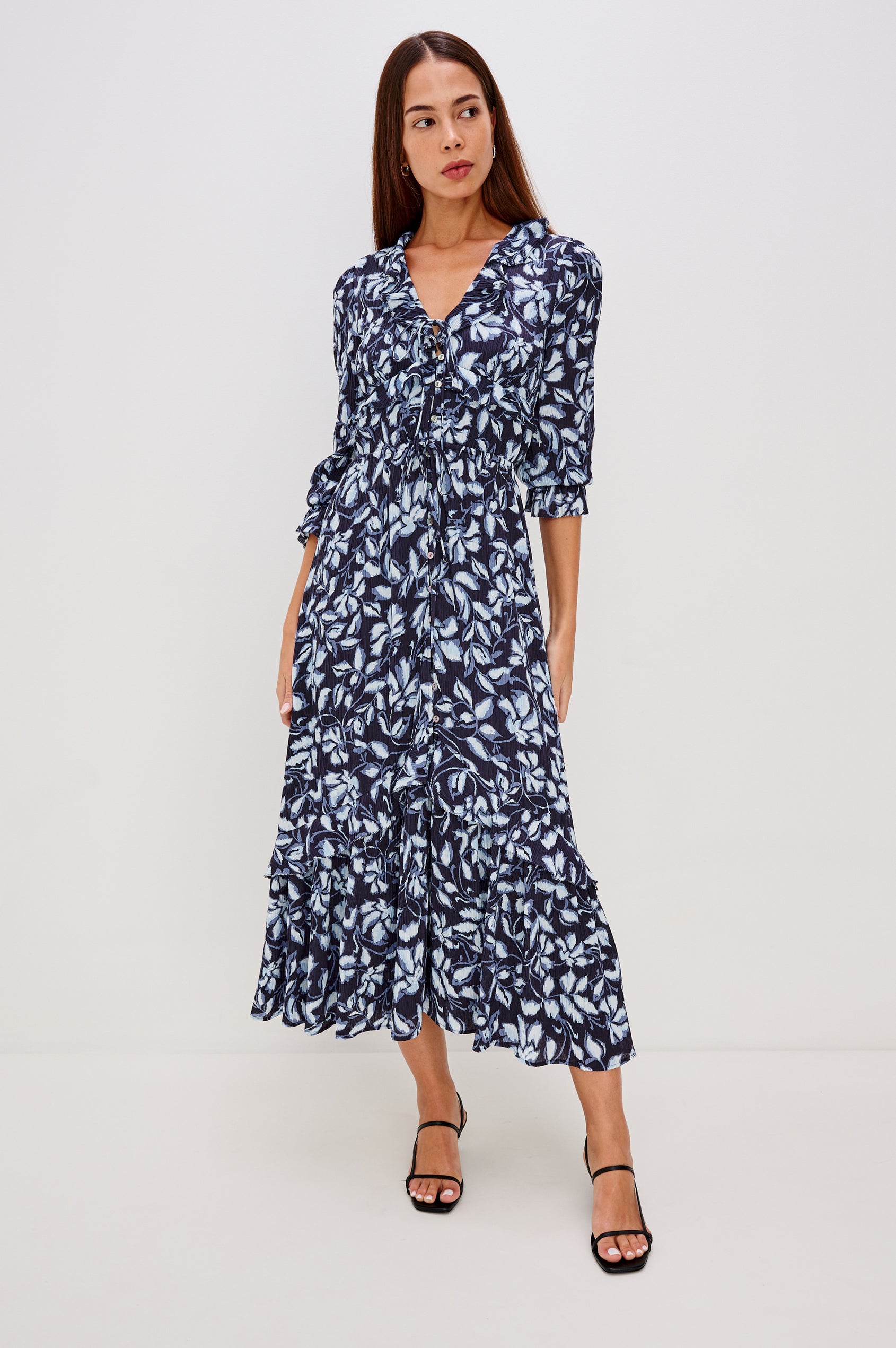 Betty is a maxi dress in pleated chiffon, it features a deep-V neckline with self ties, ruffle trim, front button-loop closures, an elastic waist with self-ties, and long sleeves with smocked cuffs in Blue Navy Jasmine Colorway.  Full body front view featured on model.