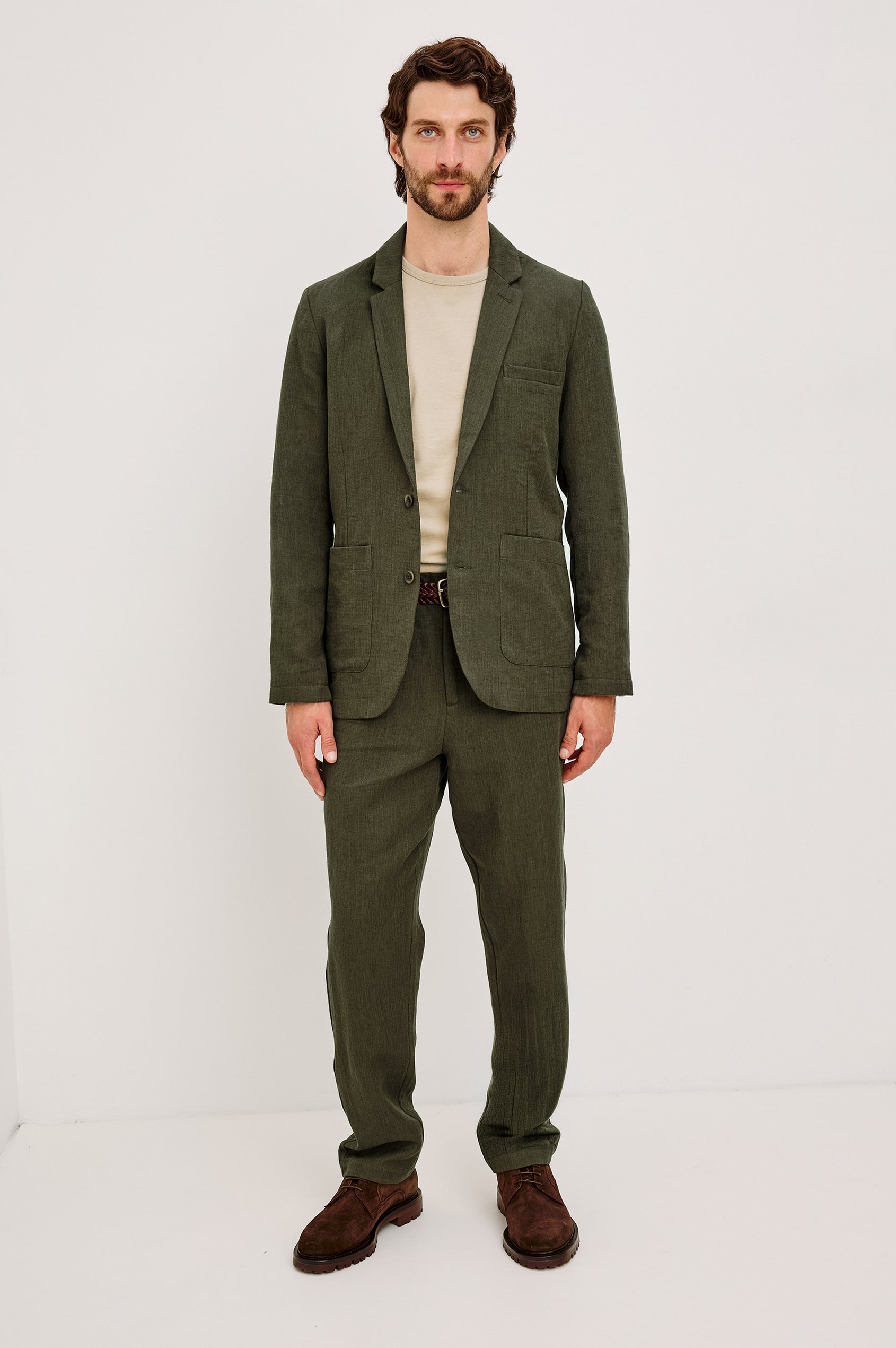 The Bond is a single breasted blazer with two buttons, one chest pocket and two patch pockets in Deep Olive colorway. Fullbody front view featured on model.