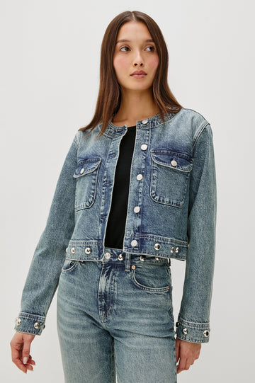 The Capistrano is a cropped denim jacket with a scoop neck, it has a button front, two chest pockets and silver studs on the sleeve cuffs and hem in Desert Sky colorway. Front view featured on model.