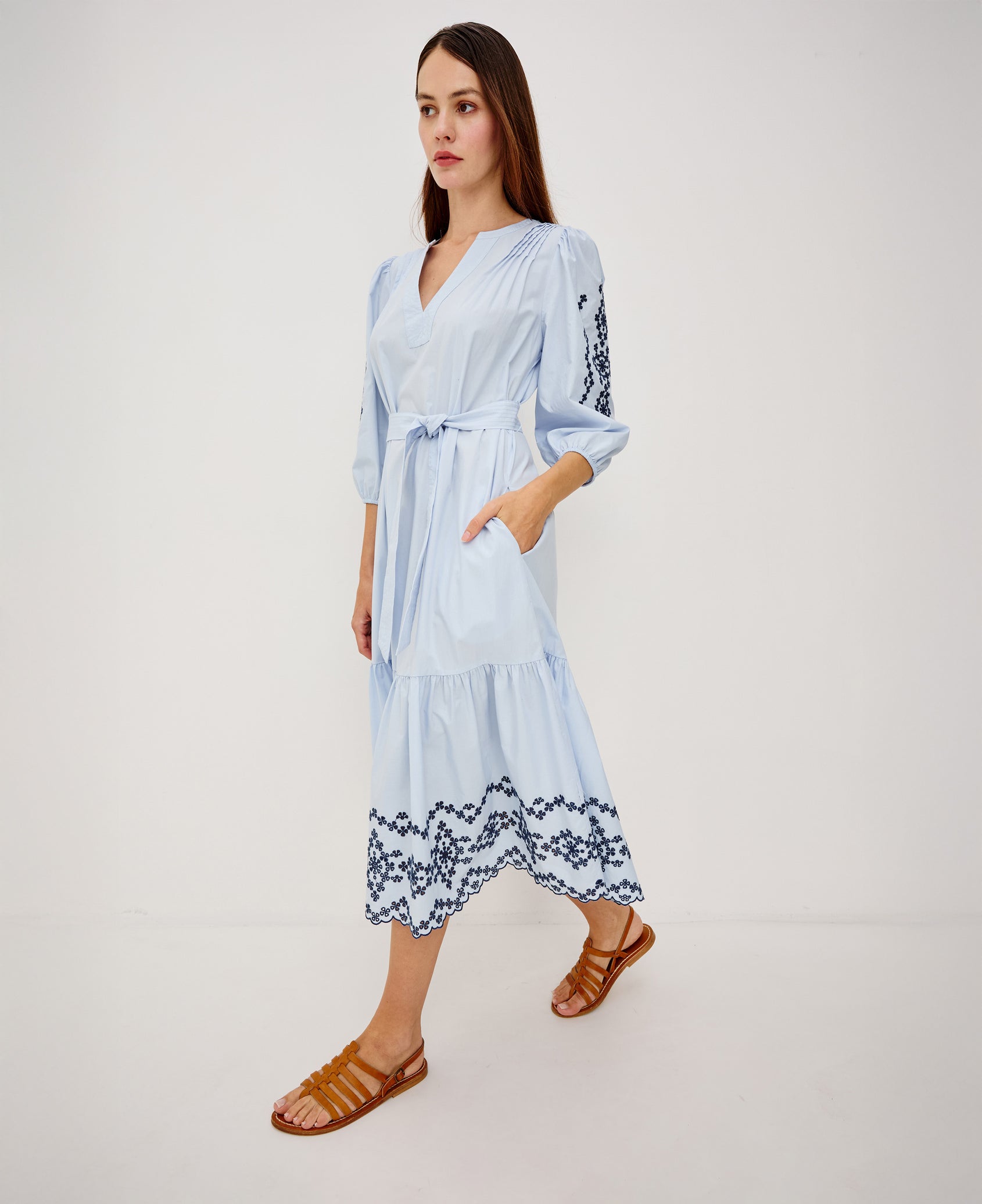 Carmel Peasant Maxi Dress in Poplin With Pleated Shoulders, V-Neckline, Three-Quarter Puff Sleeves, Adjustable Waist Ties, Side Pockets, Tiered Seam, and Contrast Embroidery at Cuffs and Hem in Blue Bird Colorway - Full Body Side View Featured on Model Walking