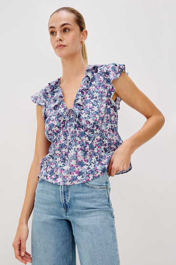 Carmine blouse, a chiffon top with ruffled cap sleeves, ruffles at the v-neckline, a front tie, and empire waist silhouette in Blue Blossom Colorway. Front View Shown on Model.
