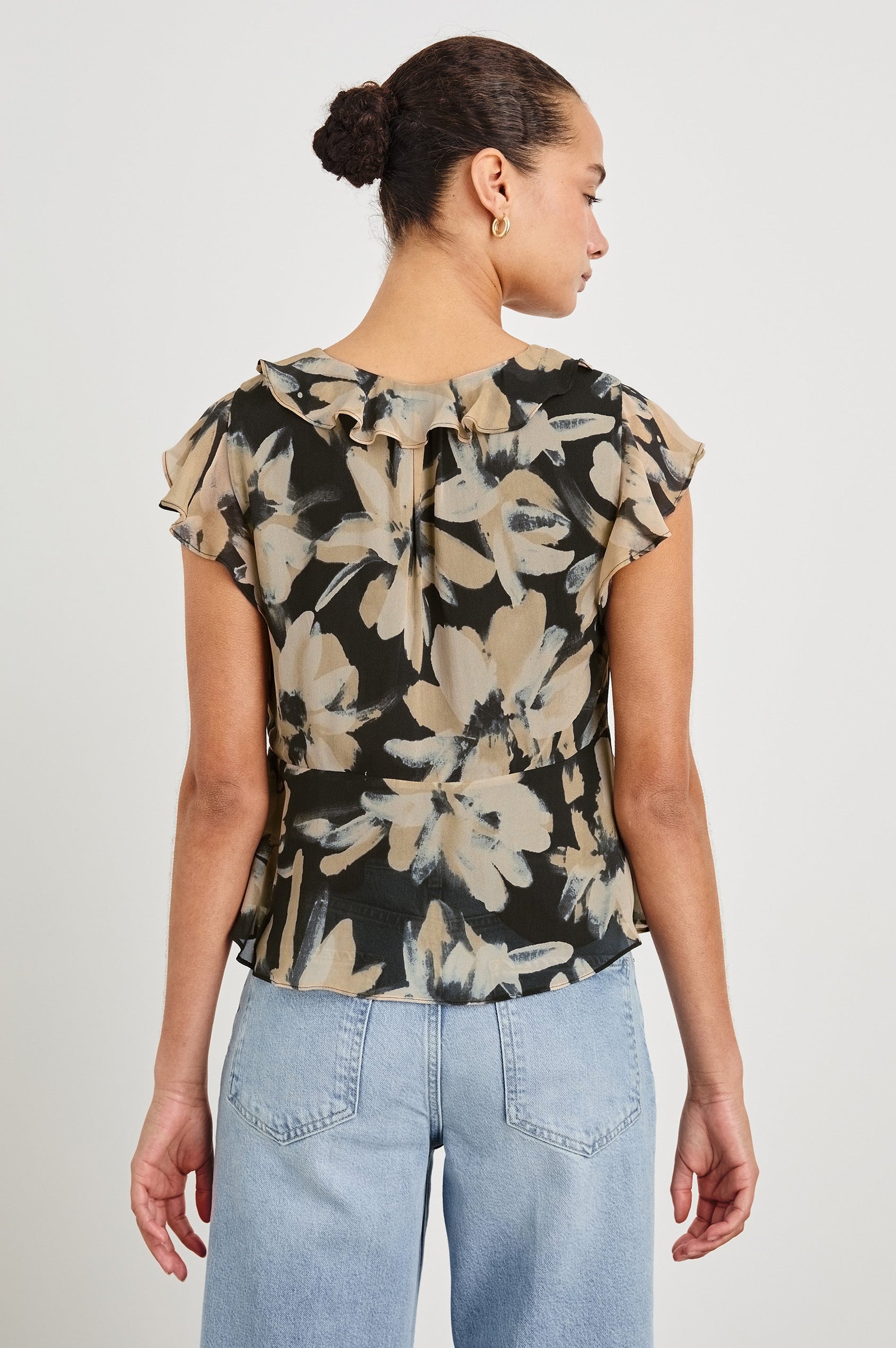 Back view of model wearing our Carmine blouse, a chiffon top with flutter sleeves, ruffles at the v-neckline, a front tie, and empire waist silhouette. Styled with light washed denim pants.