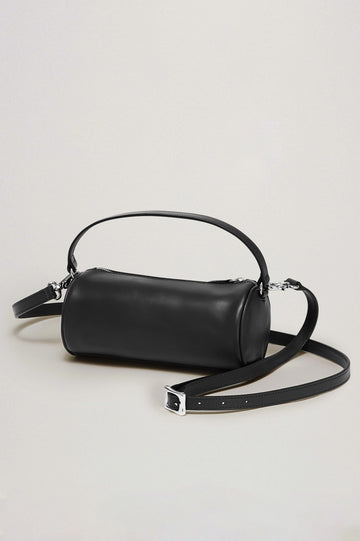 The Como is a mini barrel bag, made from smooth leather with a fixed top strap, a removable and adjustable buckled crossbody strap, a top zip, a side-embossed logo, and a branded interior label, in Black Leather colorway. Front view featured.