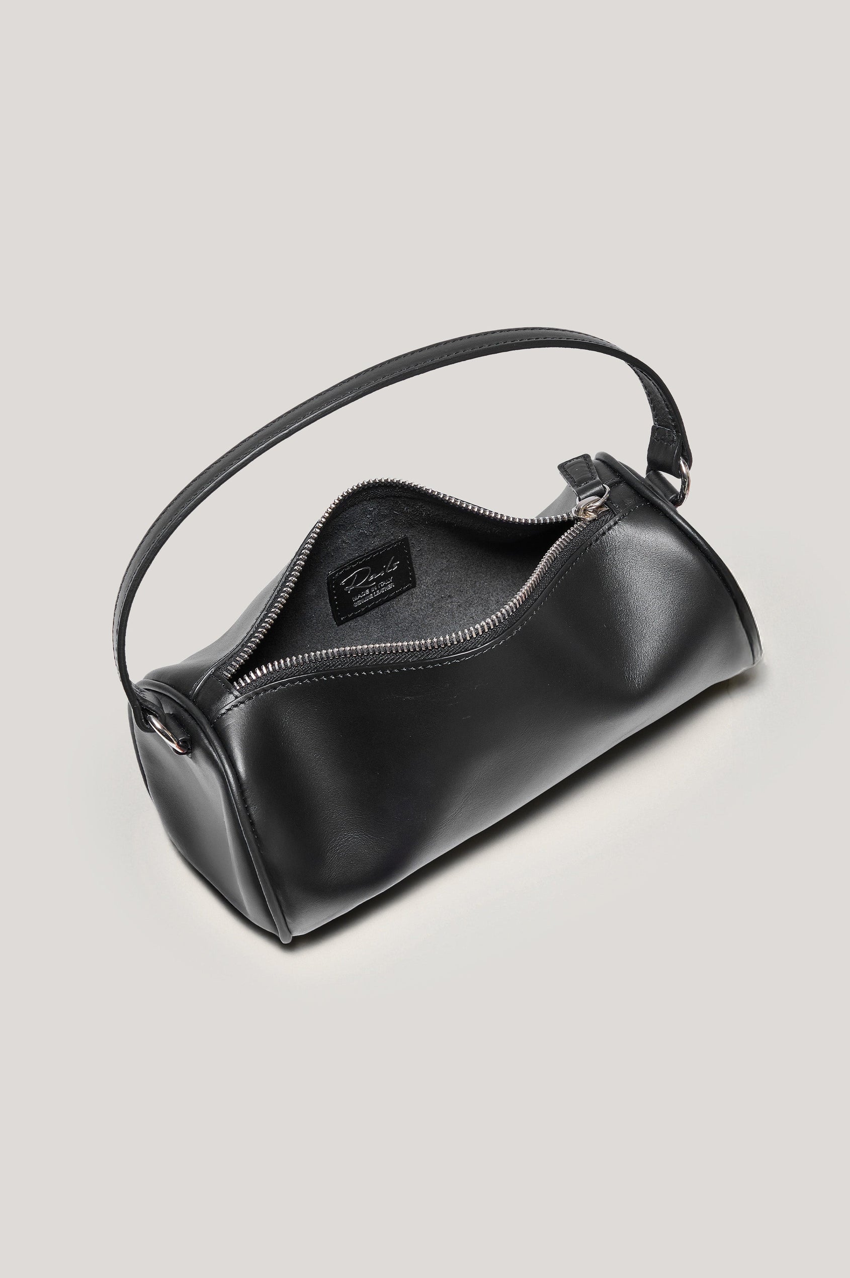 The Como is a mini barrel bag, made from smooth leather with a fixed top strap, a removable and adjustable buckled crossbody strap, a top zip, a side-embossed logo, and a branded interior label, in Black Leather colorway. Top view of interior featured.