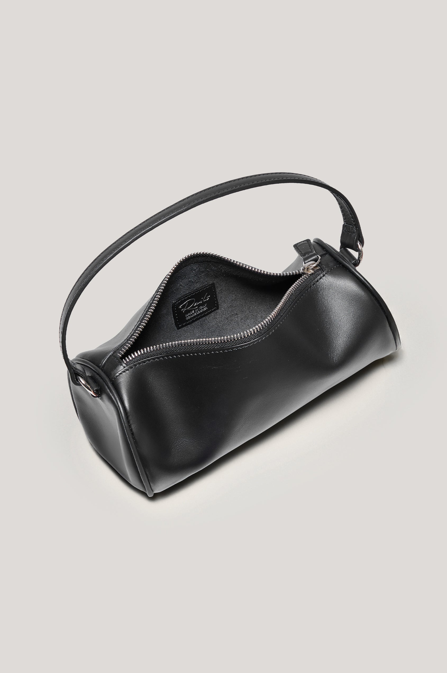 The Como is a mini barrel bag, made from smooth leather with a fixed top strap, a removable and adjustable buckled crossbody strap, a top zip, a side-embossed logo, and a branded interior label, in Black Leather colorway. Top view of interior featured.