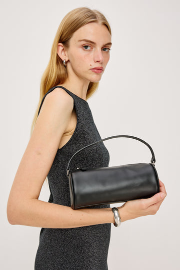 The Como is a mini barrel bag, made from smooth leather with a fixed top strap, a removable and adjustable buckled crossbody strap, a top zip, a side-embossed logo, and a branded interior label, in Black Leather colorway. Side view of model holding bag.