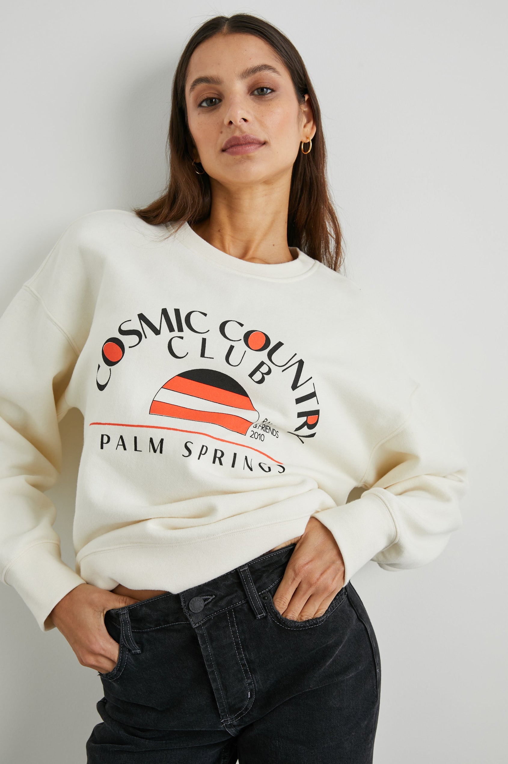 Cosmic Country Club Long-Sleeve Sweatshirt with a Crew Neck and the Front Black Text, Cosmic Country Club, Palm Springs, Rails and Friends 2010, and an Image of a Striped Setting Sun in Orange and Black in Winter White Colorway - Front View Featured on Model  
