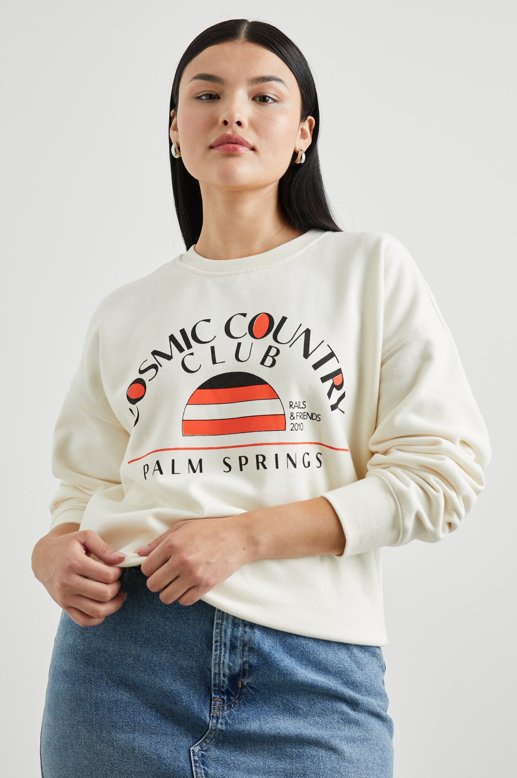 Cosmic Country Club Long-Sleeve Sweatshirt with a Crew Neck and the Front Black Text, Cosmic Country Club, Palm Springs, Rails and Friends 2010, and an Image of a Striped Setting Sun in Orange and Black in Winter White Colorway - Front View Featured on Model  