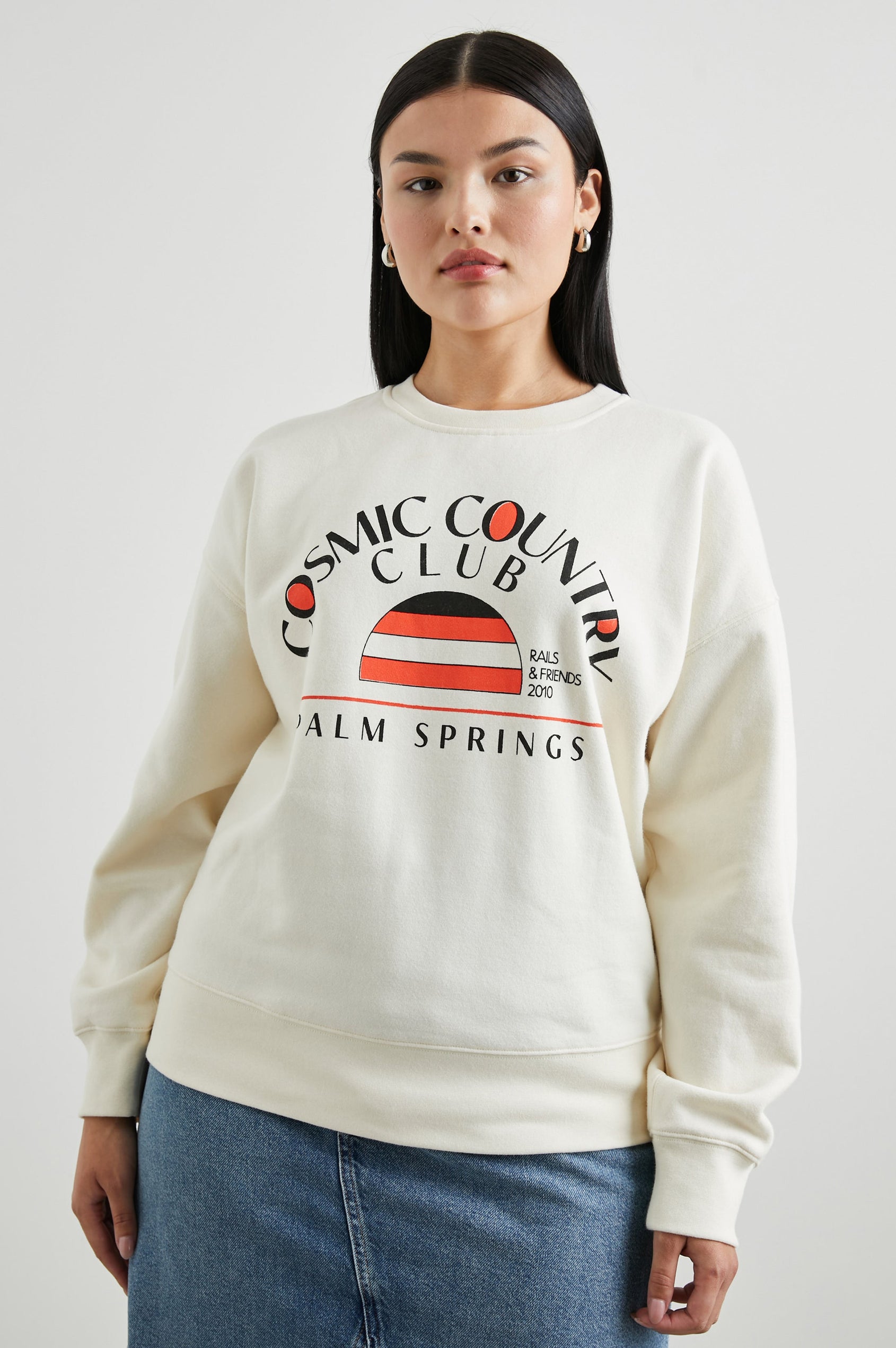 Cosmic Country Club Long-Sleeve Sweatshirt with a Crew Neck and the Front Black Text, Cosmic Country Club, Palm Springs, Rails and Friends 2010, and an Image of a Striped Setting Sun in Orange and Black in Winter White Colorway - Front View Featured on Model  