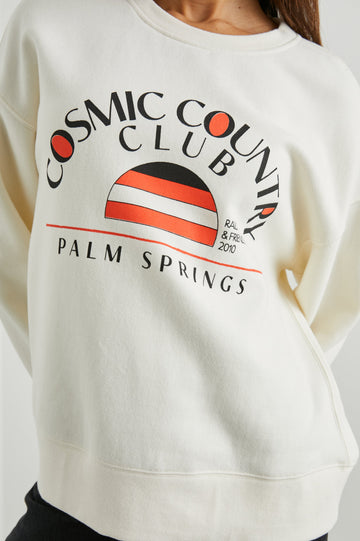 Cosmic Country Club Long-Sleeve Sweatshirt with a Crew Neck and the Front Black Text, Cosmic Country Club, Palm Springs, Rails and Friends 2010, and an Image of a Striped Setting Sun in Orange and Black in Winter White Colorway - Front View Featured on Model  