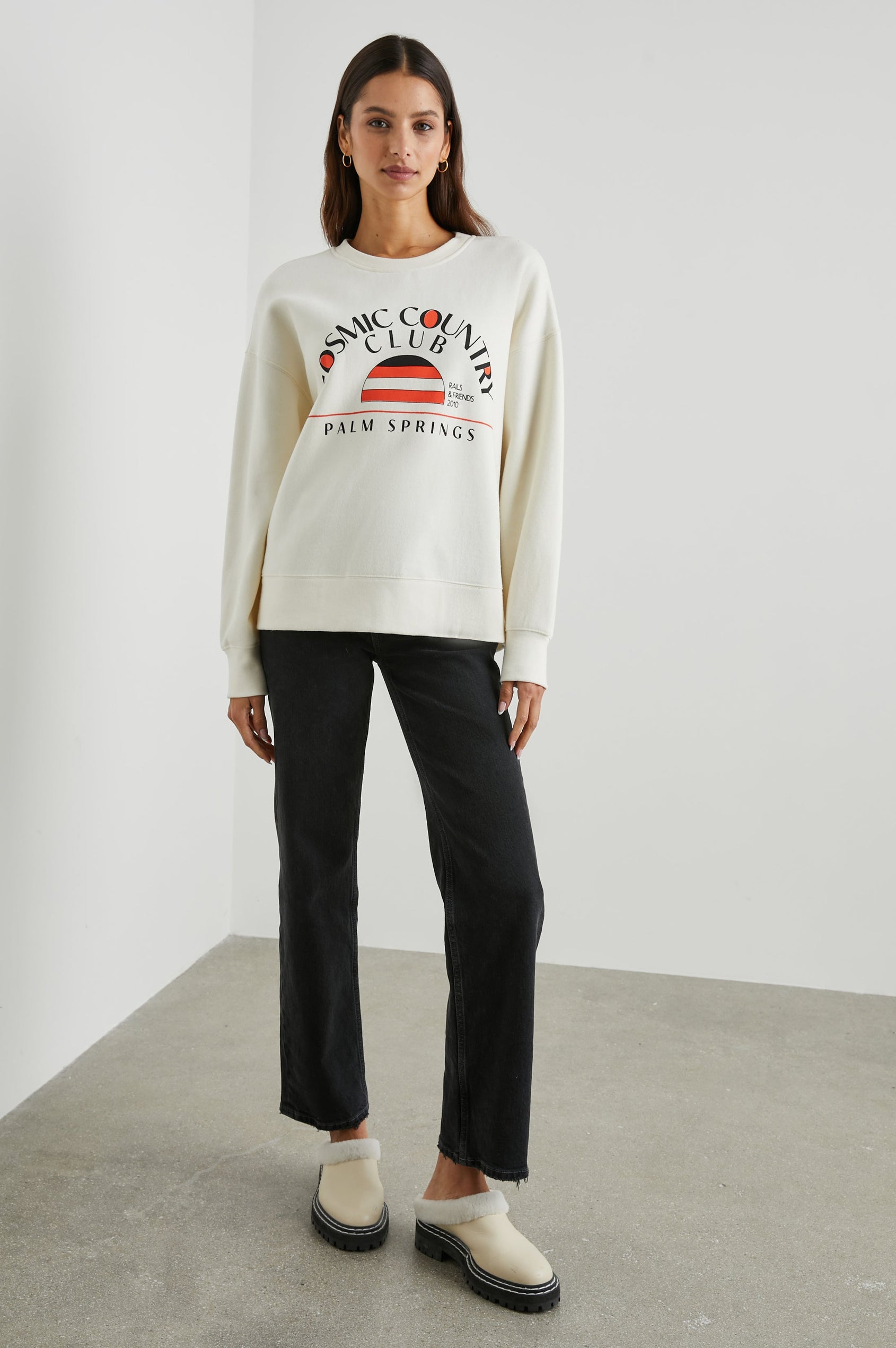 Cosmic Country Club Long-Sleeve Sweatshirt with a Crew Neck and the Front Black Text, Cosmic Country Club, Palm Springs, Rails and Friends 2010, and an Image of a Striped Setting Sun in Orange and Black in Winter White Colorway - Front Full Body View Featured on Model  