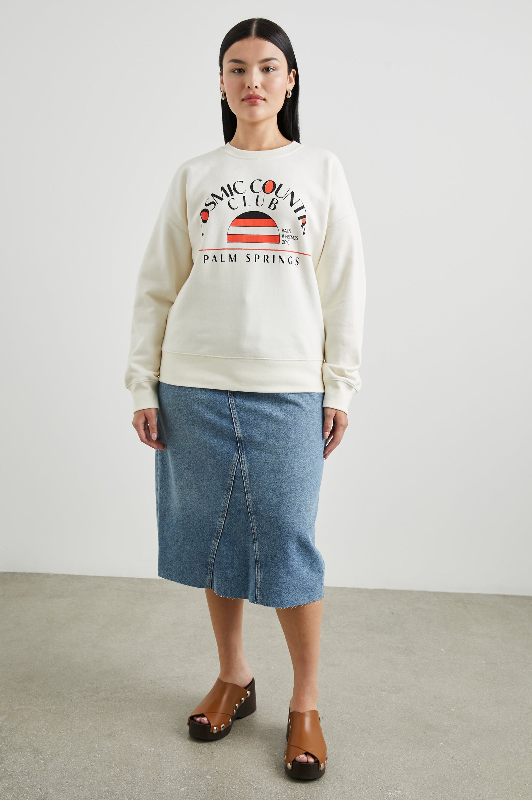 Cosmic Country Club Long-Sleeve Sweatshirt with a Crew Neck and the Front Black Text, Cosmic Country Club, Palm Springs, Rails and Friends 2010, and an Image of a Striped Setting Sun in Orange and Black in Winter White Colorway - Front Full Body View Featured on Model  