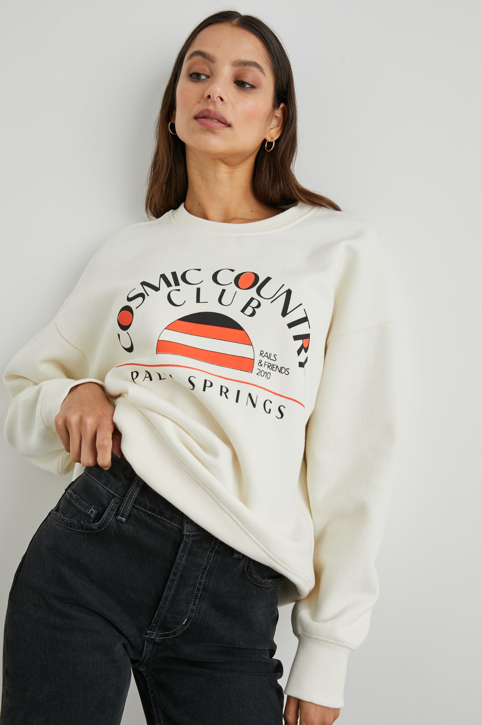 Cosmic Country Club Long-Sleeve Sweatshirt with a Crew Neck and the Front Black Text, Cosmic Country Club, Palm Springs, Rails and Friends 2010, and an Image of a Striped Setting Sun in Orange and Black in Winter White Colorway - Front View Featured on Model  