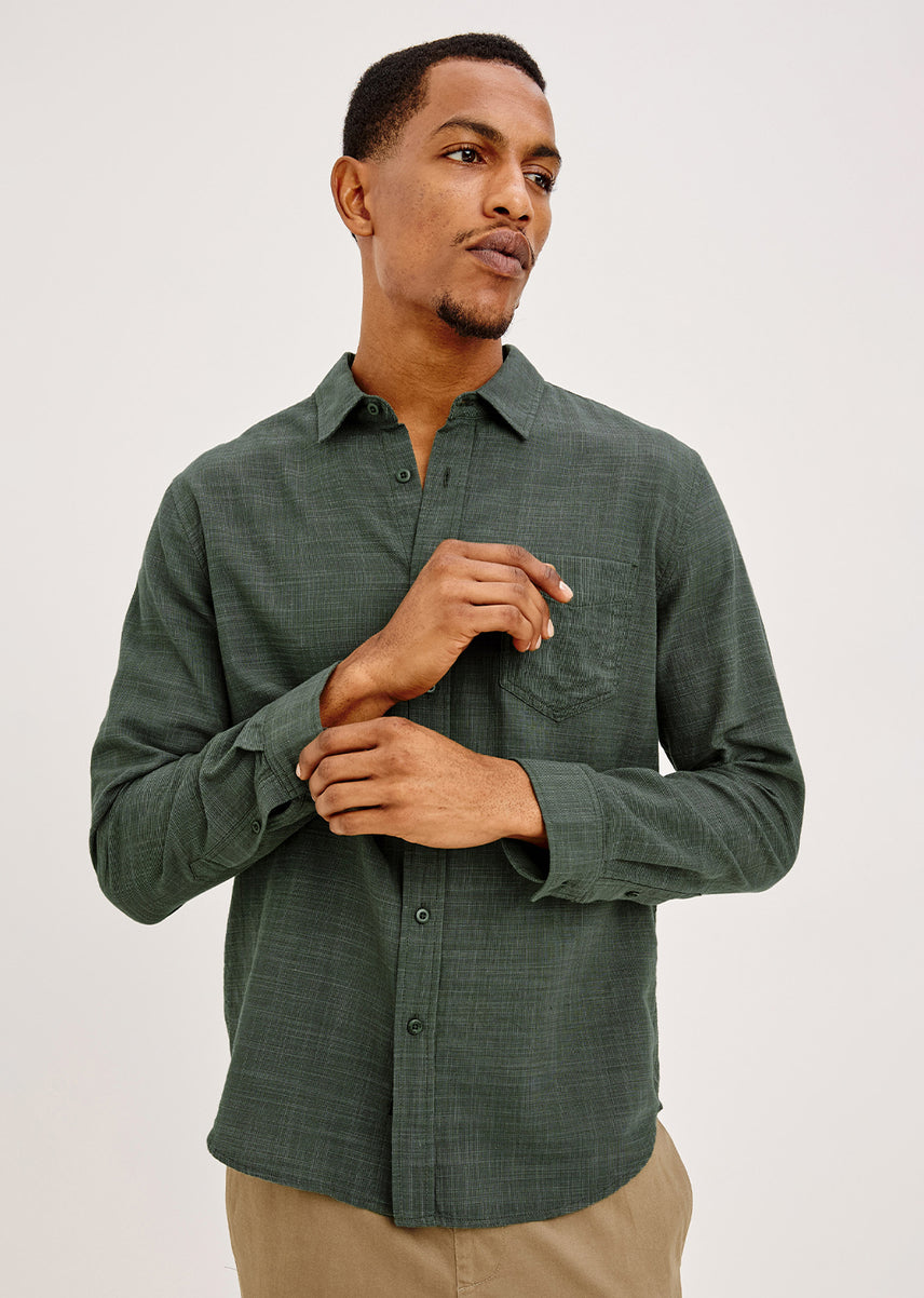 A man wearing a dark green long-sleeve button-down shirt with a subtle textured pattern, paired with khaki pants, photographed against a light gray studio background.