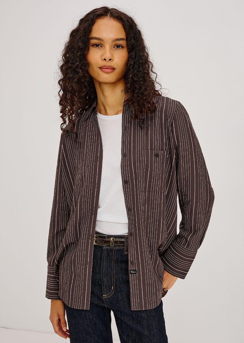 A woman with long curly dark hair models a brown and dark gray vertically striped button-up shirt jacket over a white t-shirt and dark blue jeans, photographed against a light gray studio background.