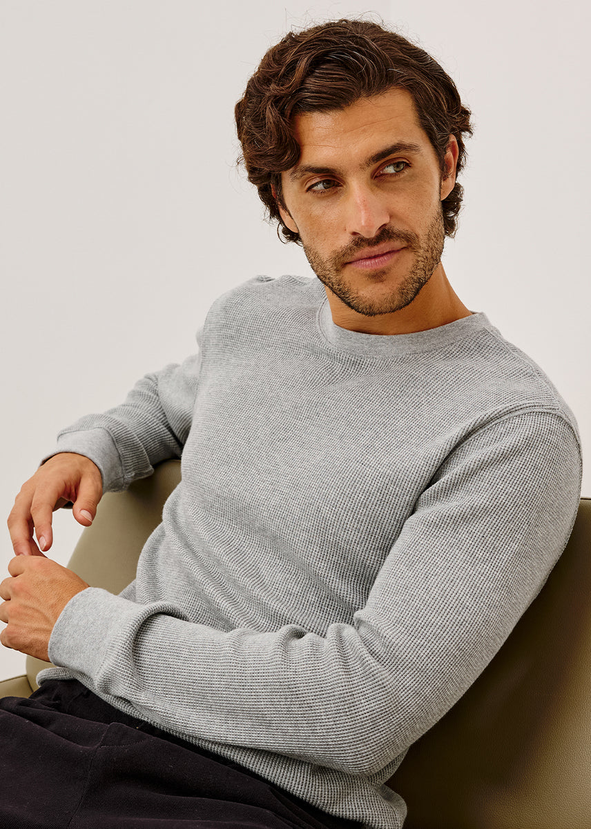 A man wearing a light gray ribbed knit crewneck sweater, seated against a neutral light gray background.
