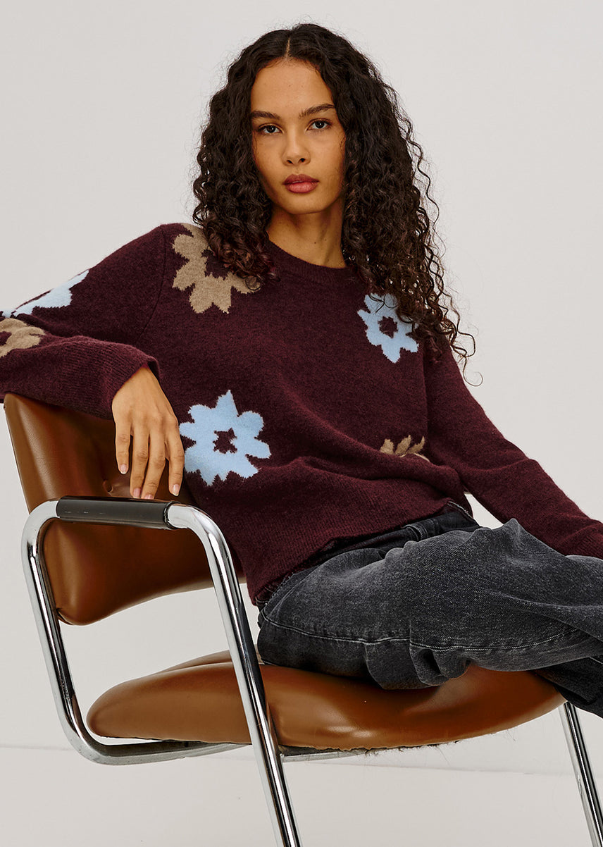 A woman with long curly dark hair sits on a brown leather chair wearing a fuzzy crewneck sweater in deep burgundy with light blue and beige floral patterns, paired with dark gray jeans.