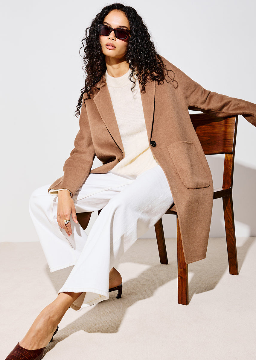 A woman models a long camel brown wool-blend coat over a cream top and white wide-leg trousers, seated on a wooden chair against a light beige studio background.