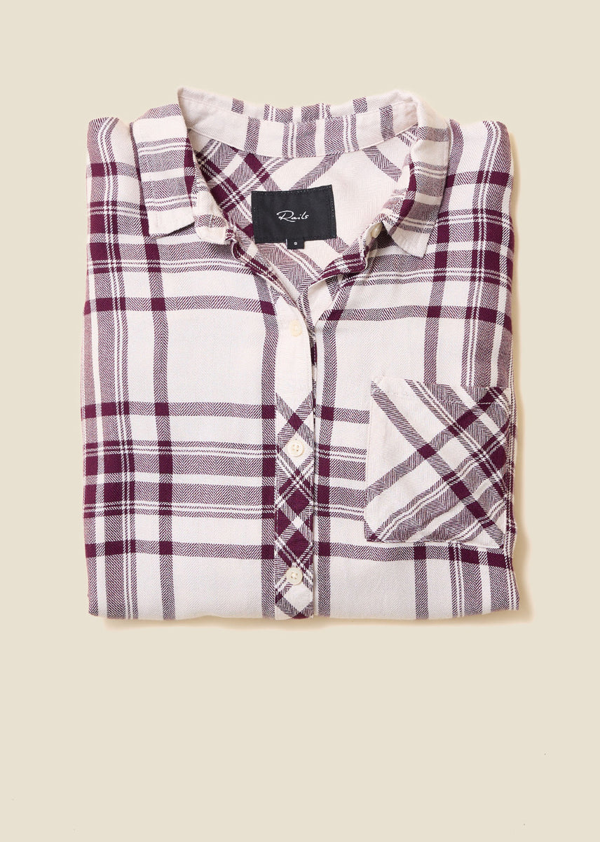 Folded cream flannel shirt with a burgundy plaid pattern and chest pocket, with a Rails label at the collar, on a beige background.