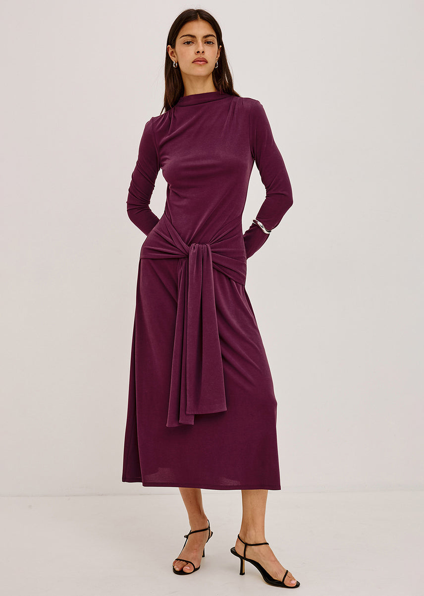 Woman in a plum long-sleeve midi dress with a draped waist tie and black strappy heeled sandals.